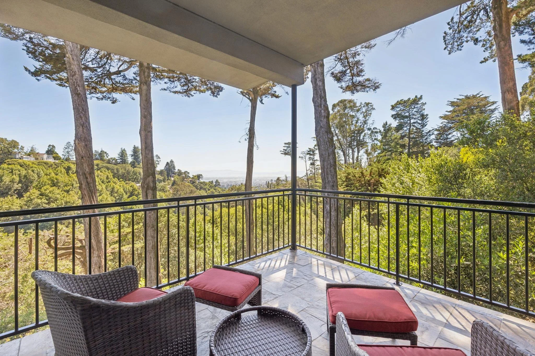 A balcony with wicker chairs and red cushions overlooking a lush green forest, with tall trees and a view of distant hills or water.