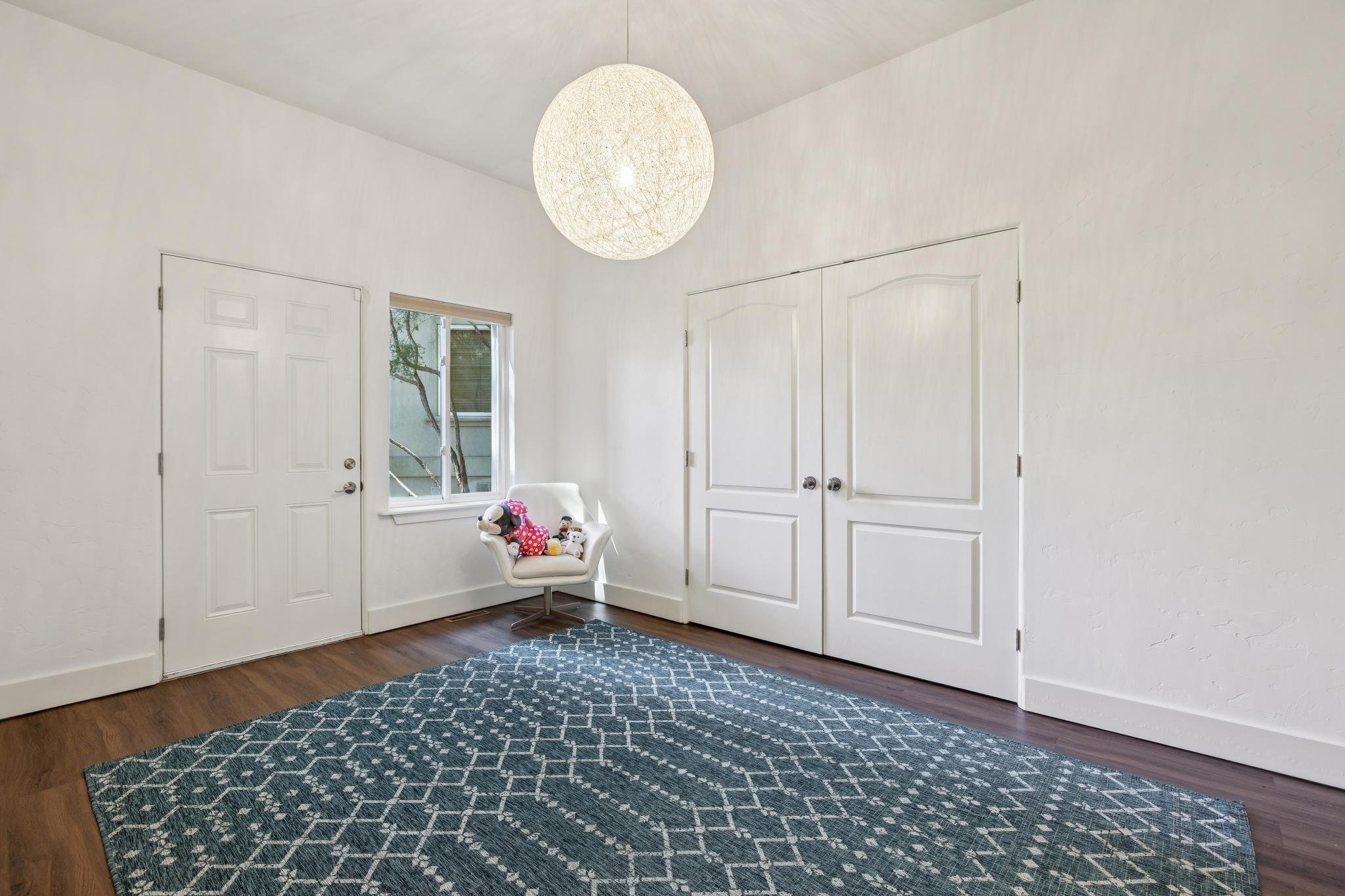 A white chair with stuffed animals in a room corner near a window, with white walls, a patterned gray rug, and a ceiling light fixture.