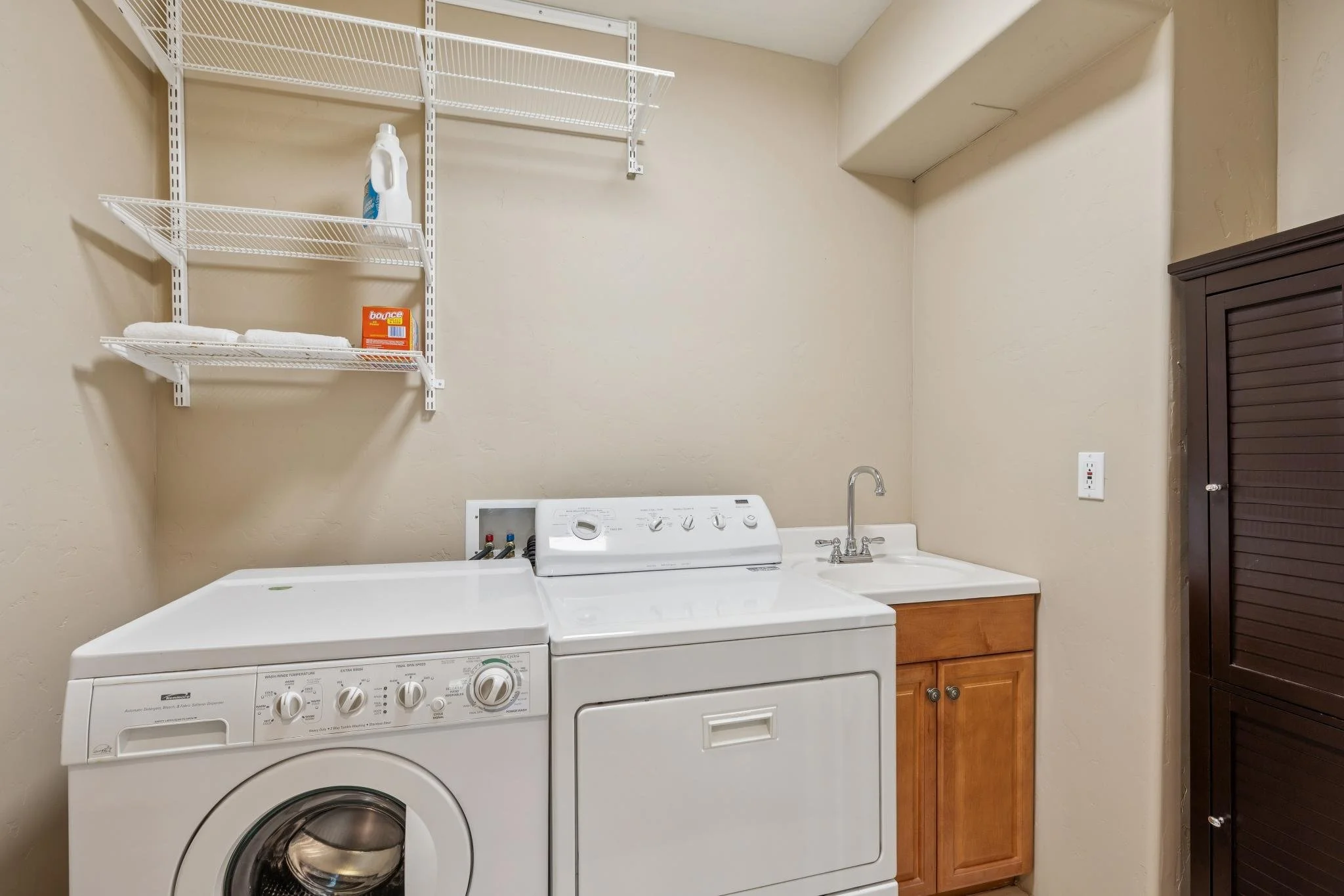 A laundry room with a white washing machine and dryer side by side, a wooden cabinet on the right, wire shelves above holding laundry supplies, a small sink with a faucet between the appliances, and an electrical outlet on the wall.