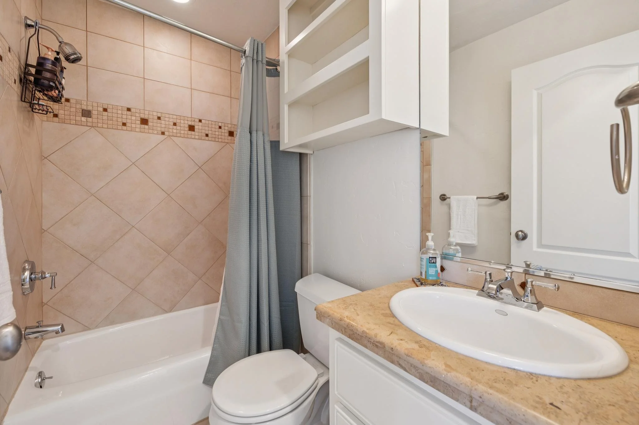A bathroom with a shower area enclosed by a shower curtain, a toilet, a sink with a beige countertop, a mirror with a cabinet above, and toiletries including hand soap and sanitizer on the counter.