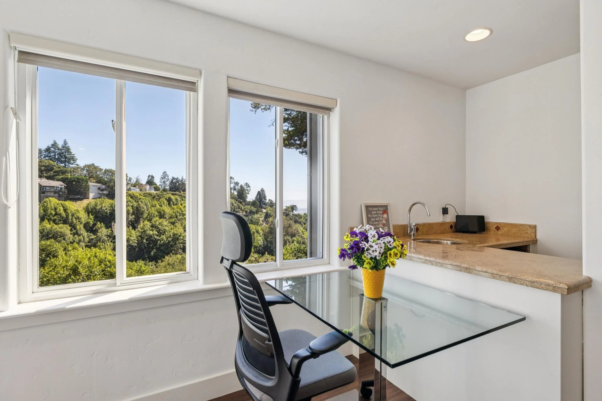 Interior view of a bright room with large windows showing a scenic green landscape and trees outside. There is a glass table with a black office chair and a yellow vase with purple and white flowers on the table. A framed quote is leaning against the