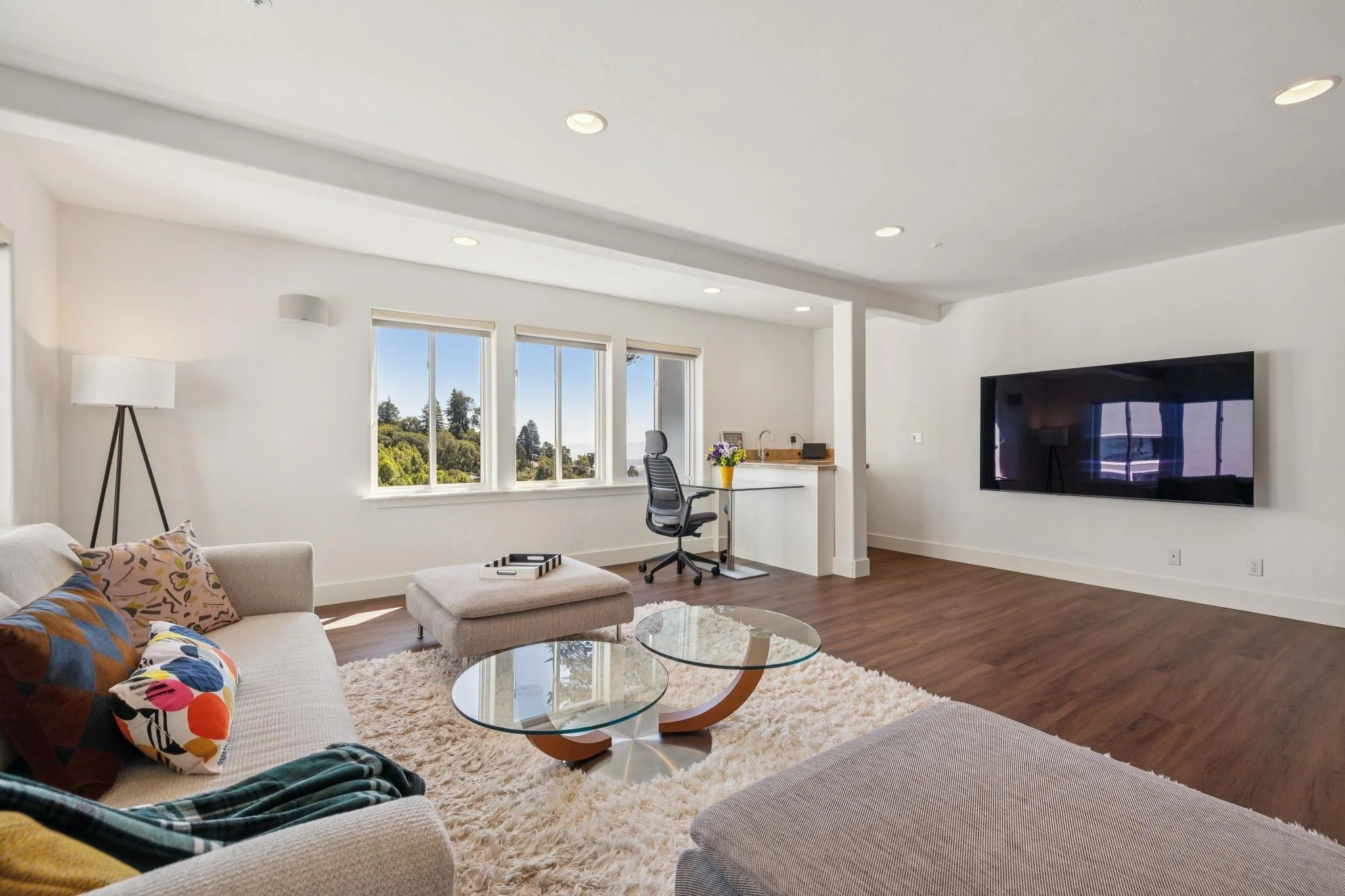 Living room with white walls, large windows showing trees outside, a beige sofa with colorful pillows, a glass coffee table, a shaggy white rug, a black office chair near a white desk with flowers, a large flat-screen TV, a white floor lamp, recessed