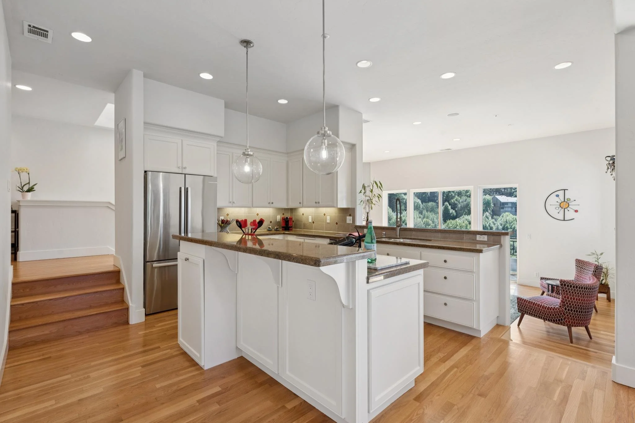 Open-concept kitchen with white cabinetry, a large granite island, pendant lights, stainless steel refrigerator, and large windows overlooking trees.
