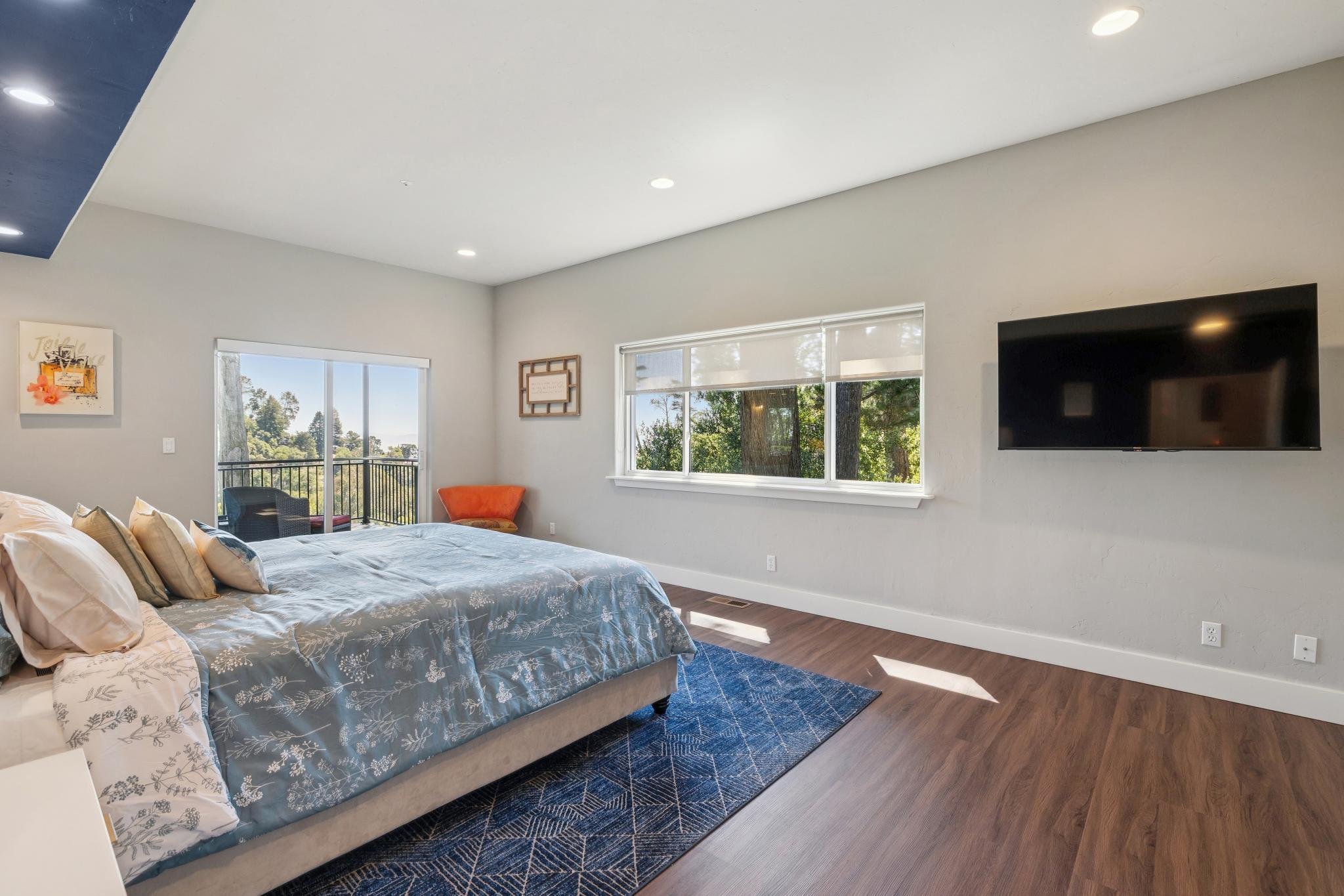 Modern bedroom with a bed, decorative pillows, a blue area rug, a flat-screen TV on the wall, two large windows with views of trees outside, and a small balcony with outdoor furniture.