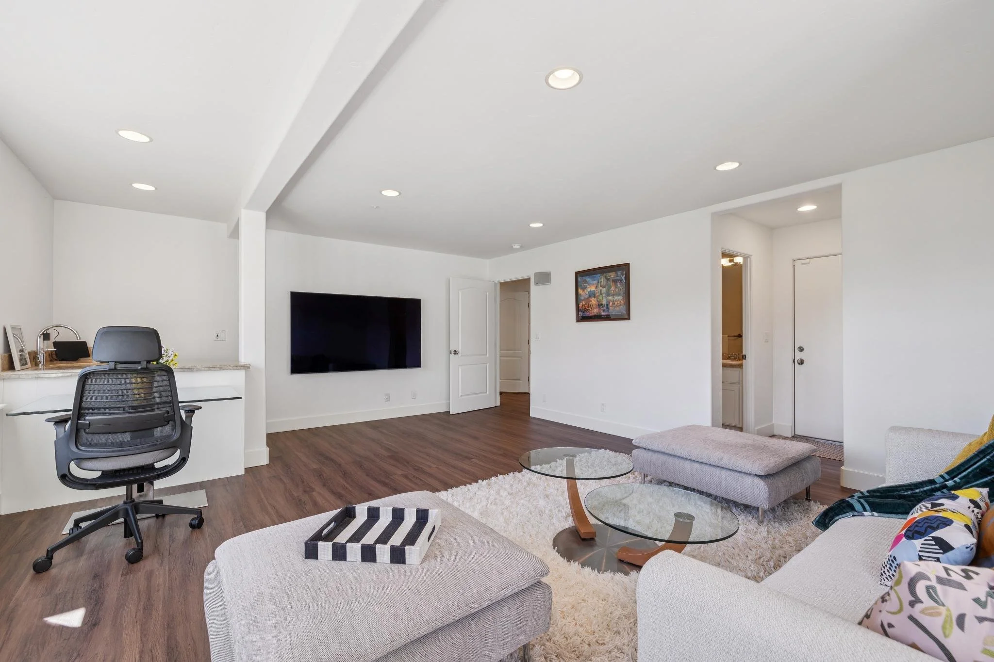 Living room with white walls, hardwood flooring, a large flat-screen TV, a white couch, a gray ottoman, glass coffee tables, a desk and chair, colorful pillows, a framed painting, and ceiling lights.