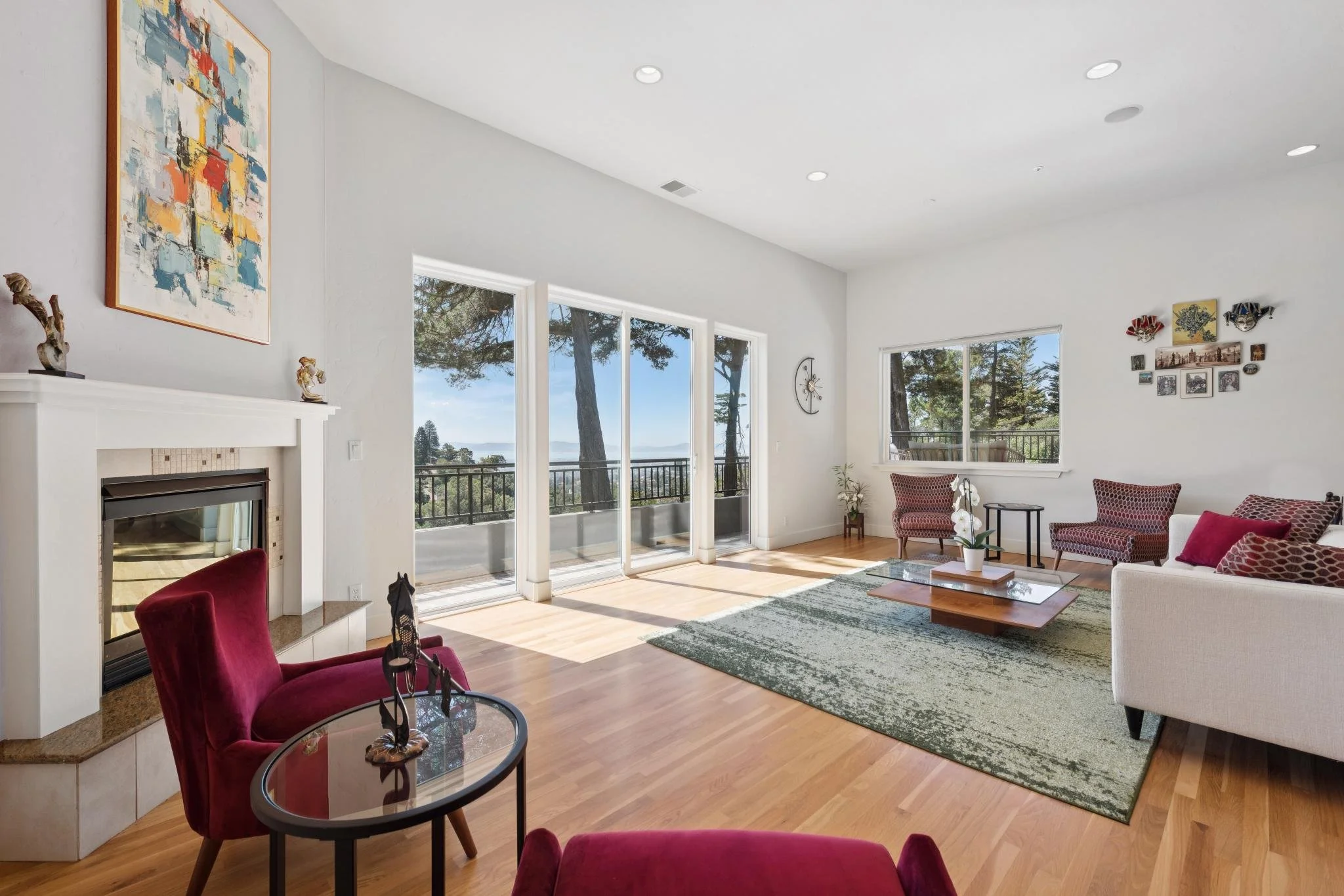 Living room with large windows showing trees and a view outside, a fireplace, red velvet chairs, a beige sofa with red pillows, a glass coffee table with a orchid plant, artwork on the walls, and wooden flooring.