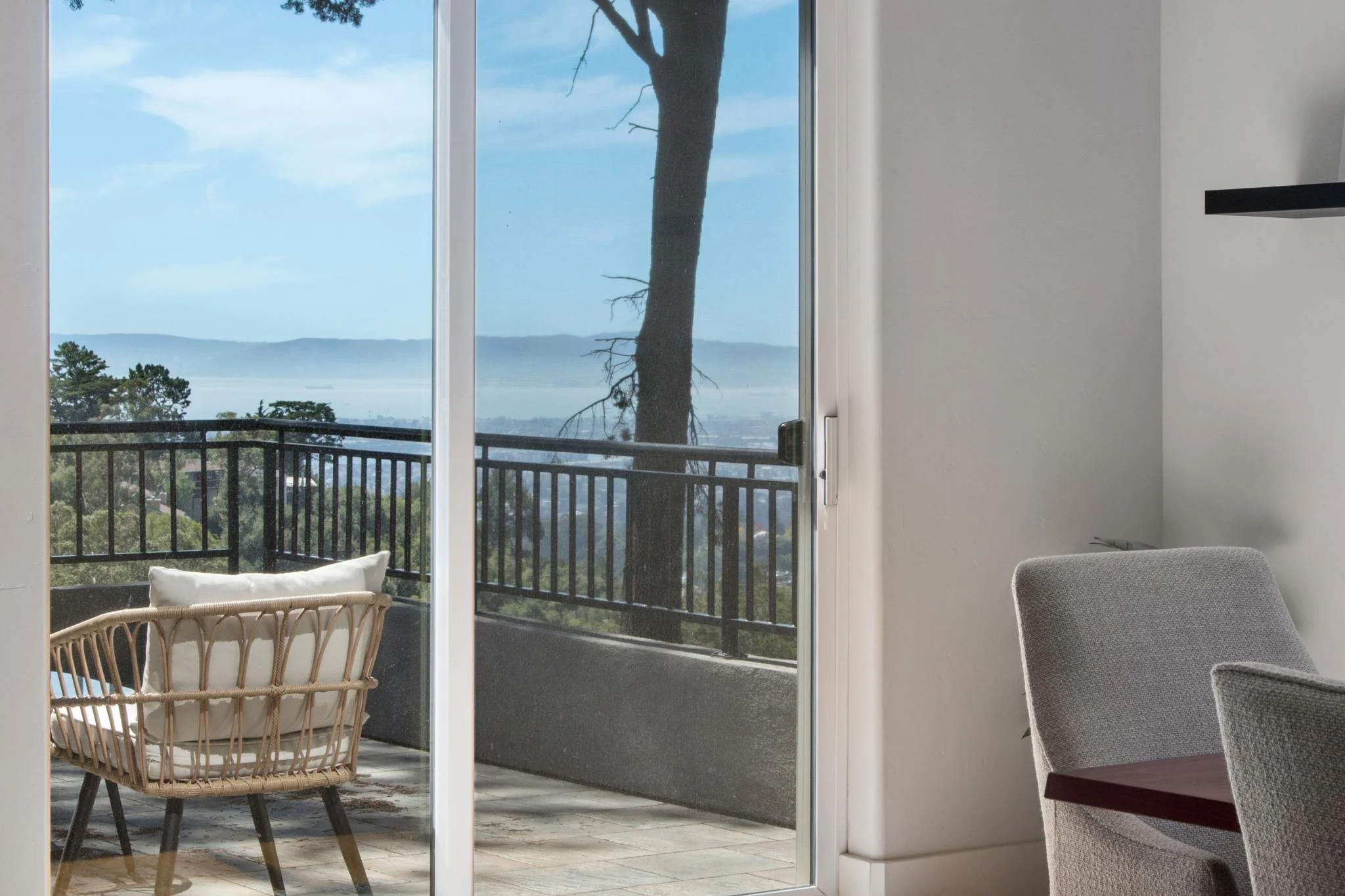 A view of a balcony through a glass sliding door, with a landscape featuring mountains, trees, and a clear sky in the background. Inside, there is a wicker chair with a cushion and a gray upholstered chair partially visible.