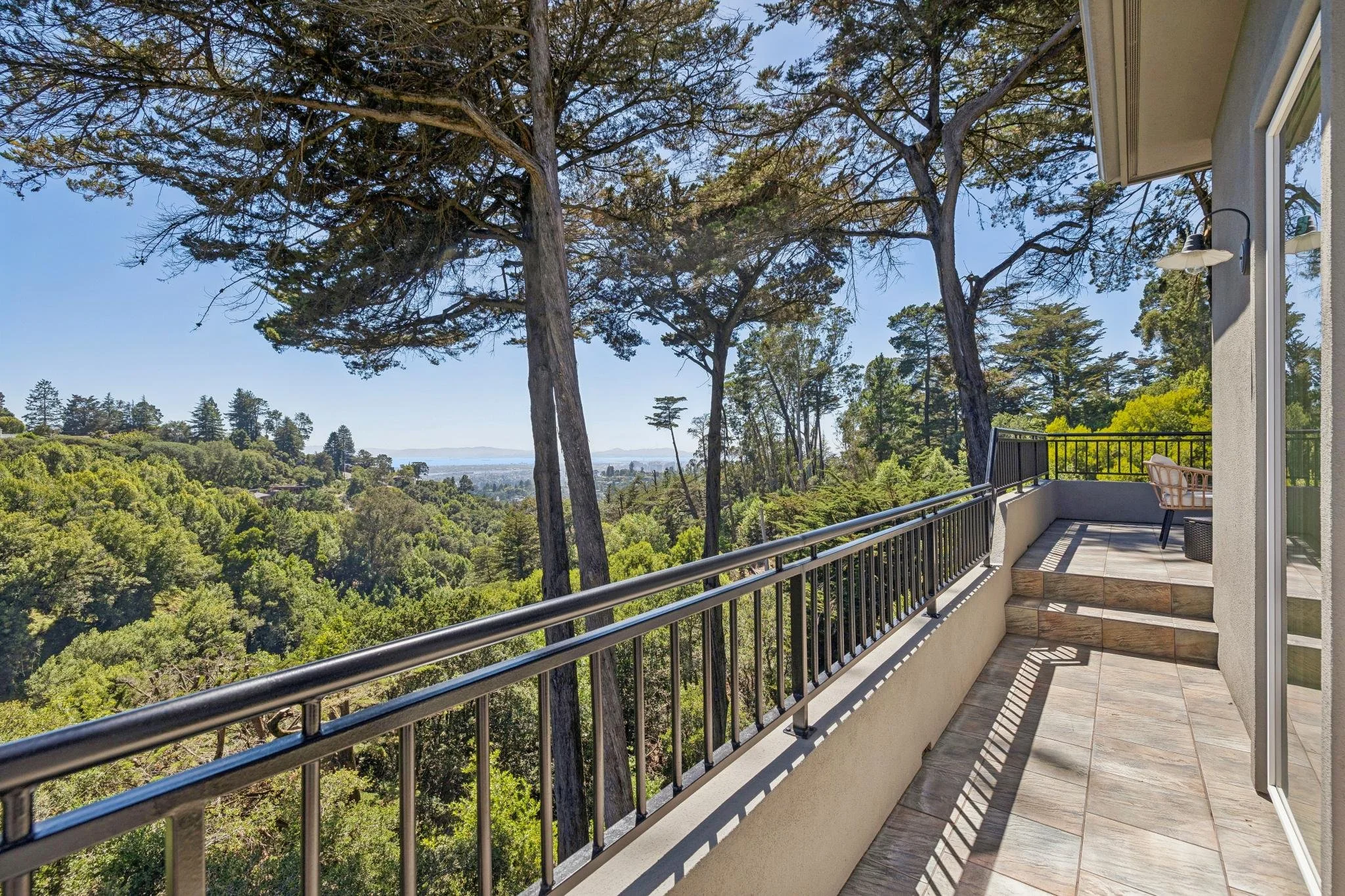 Balcony with metal railing overlooking a green forested landscape with trees and distant hills under a clear blue sky.
