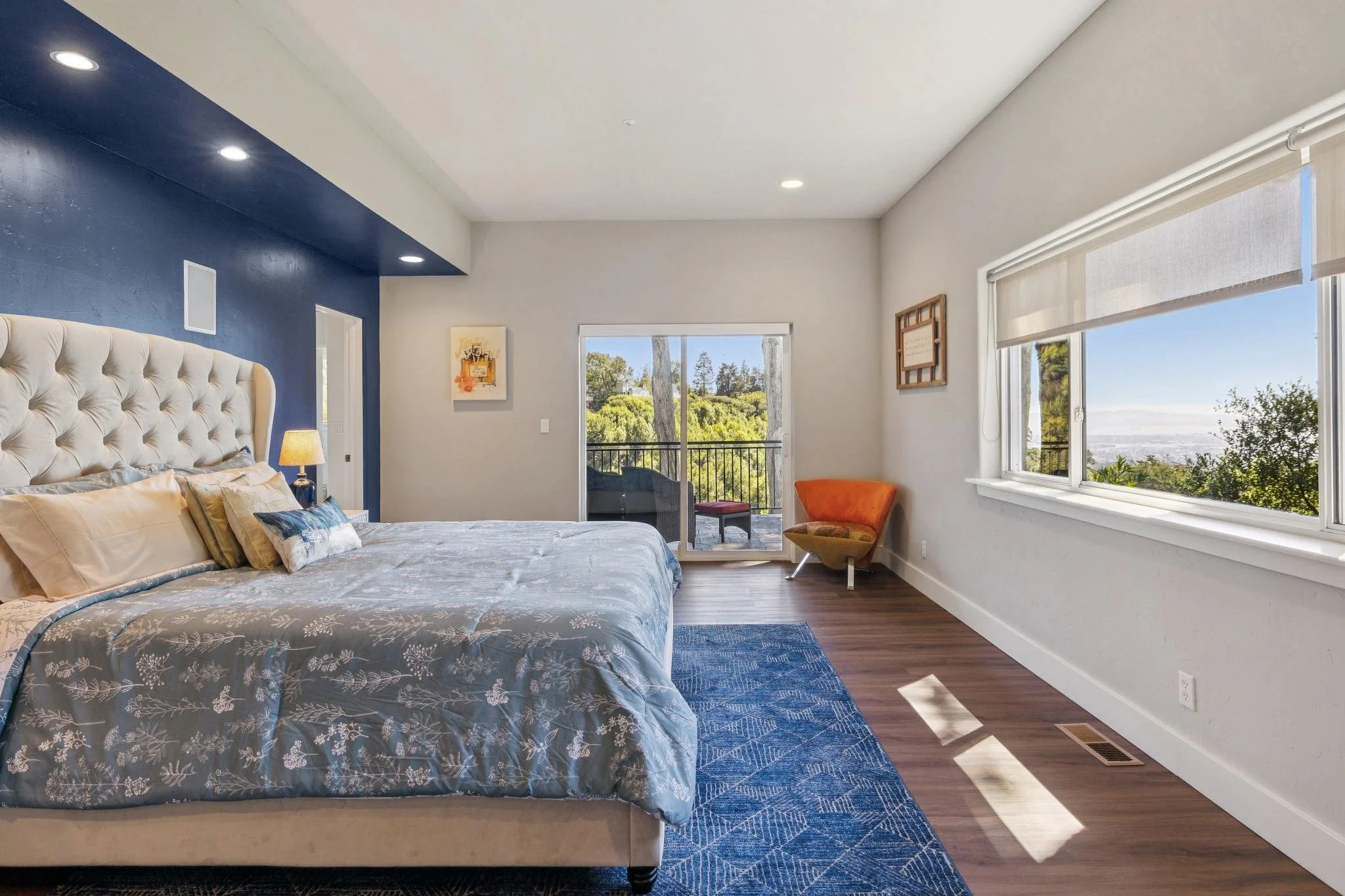 A bedroom with a large bed covered with floral-patterned bedding, a tufted cream-colored headboard, a side table with a lamp, and a blue accent wall. Windows with blinds reveal an outdoor view with trees and a clear sky.