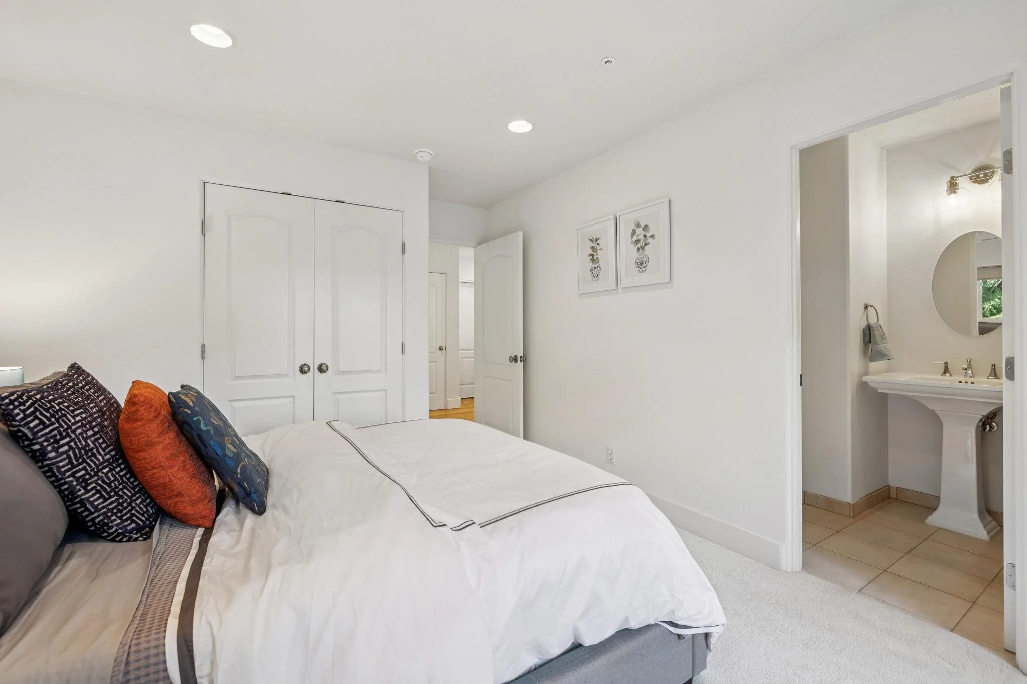 A bedroom with a white ceiling and walls, a bed with white and brown linens, and several decorative pillows in black, burnt orange, and blue patterns. There are double closet doors on the left and an open doorway on the right leading to a bathroom wi