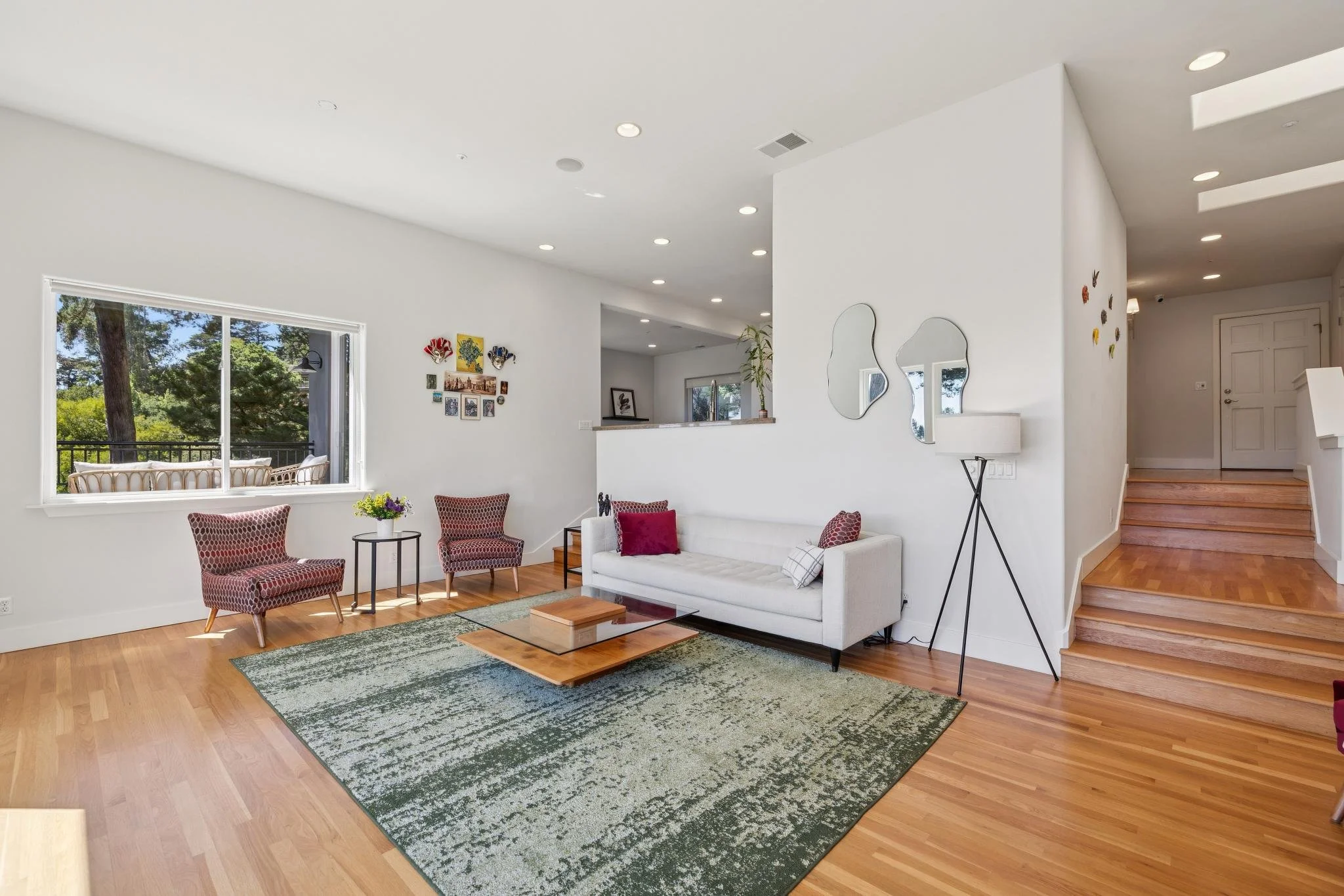 Living room with white walls, large window showing trees outside, wooden floors, patterned chairs, white sofa, wall mirrors, and decorative wall art.