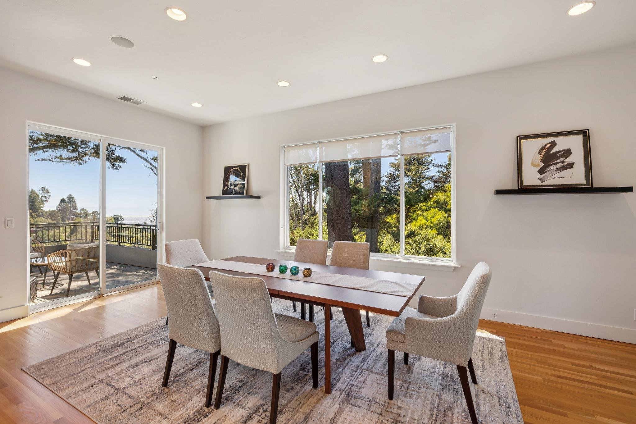 A bright dining room with a wooden table and six beige upholstered chairs. Large windows with white blinds showcase a view of trees outside. Two black floating shelves with framed abstract art hang on the white walls. There is a sliding glass door le