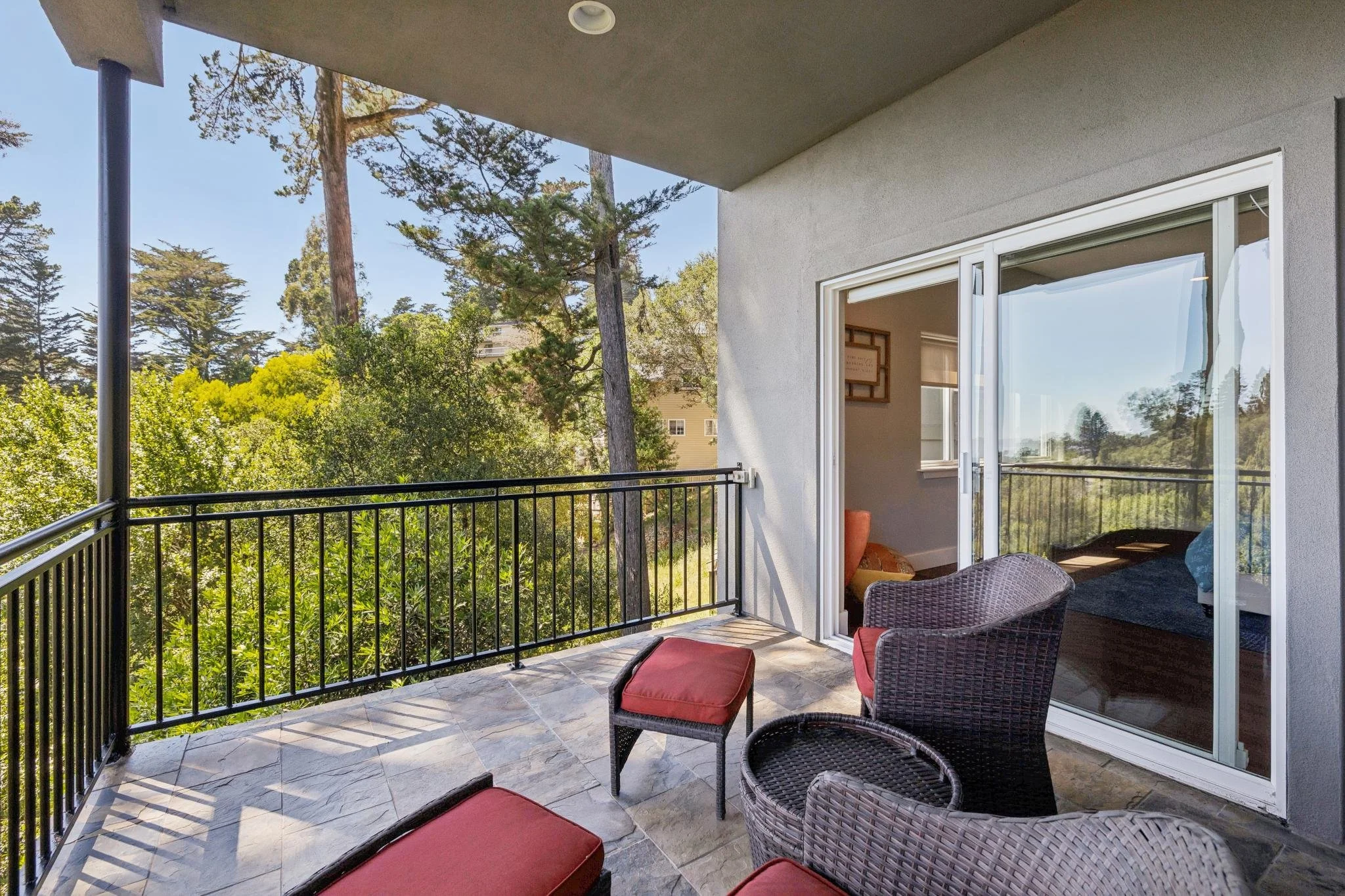 Balcony with outdoor furniture, including chairs with red cushions, overlooking lush green trees and a neighboring building, with sliding glass doors leading inside.