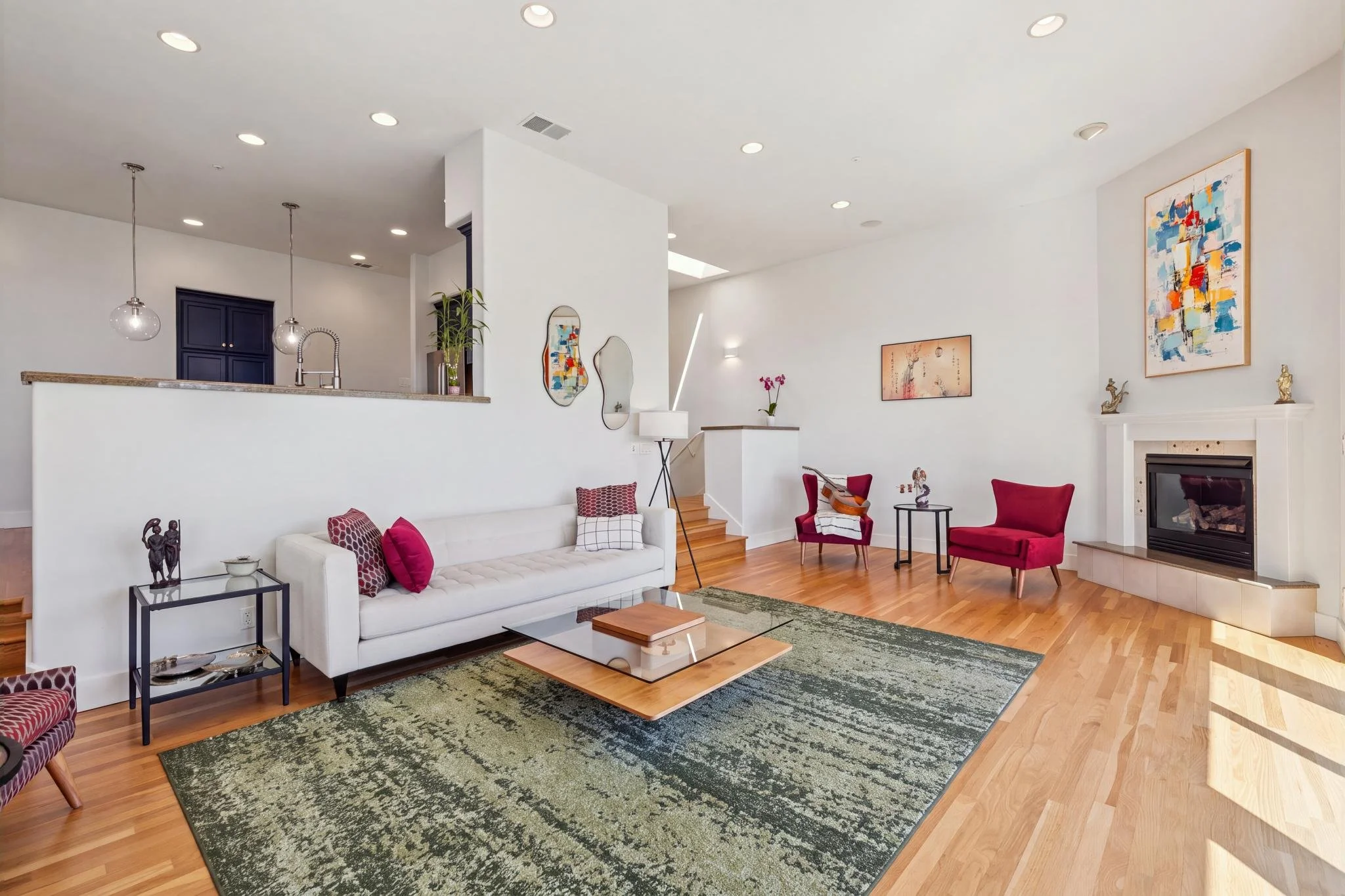 Living room with white walls, hardwood flooring, a white sofa with red and black pillows, two red armchairs, a dark metal side table with two sculptures, a fireplace with decorative items on the mantel, colorful abstract artwork on the walls, a green