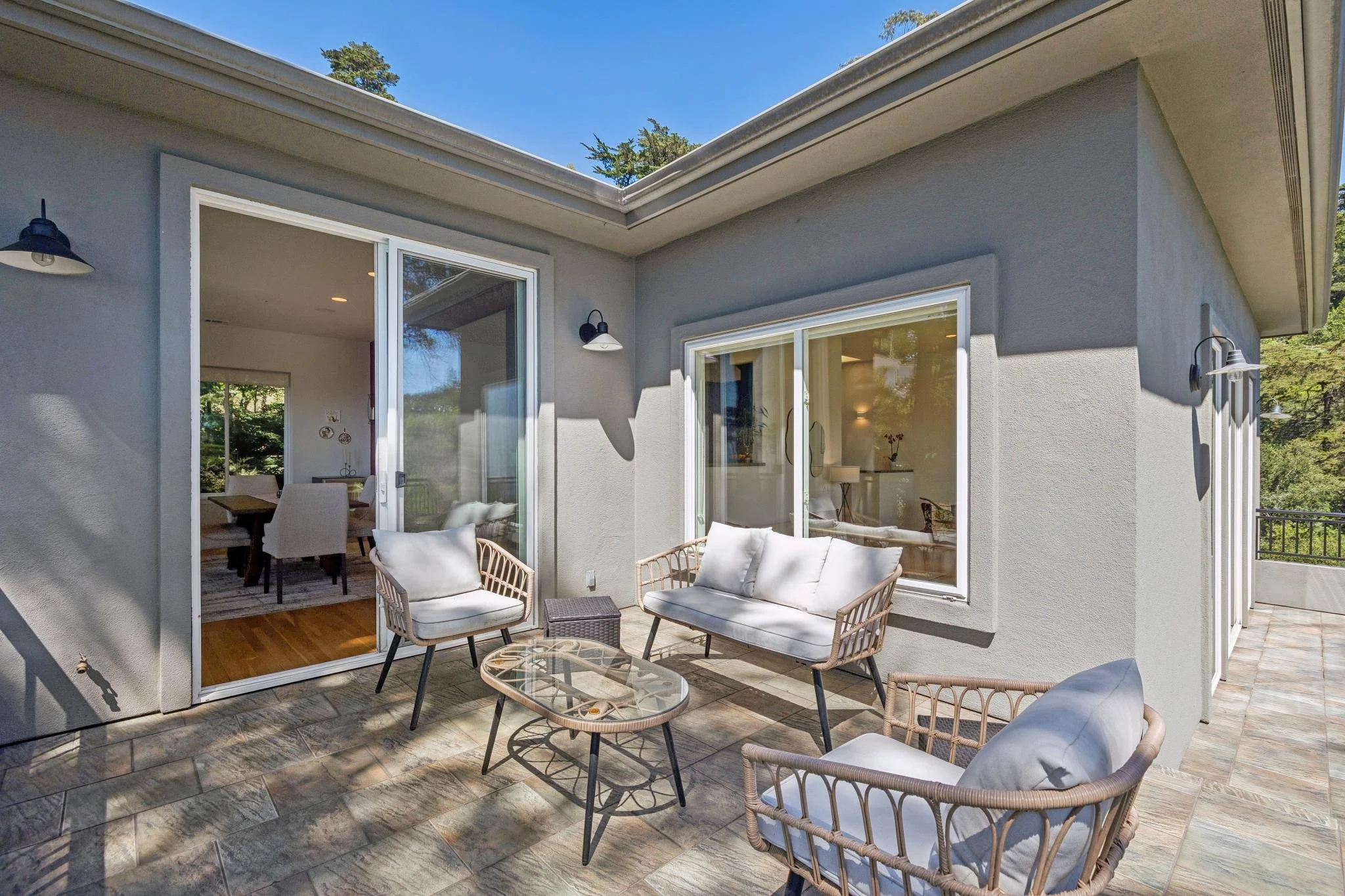 An outdoor patio area with wicker chairs and glass tables, adjacent to a house with sliding glass doors and windows. The patio is partially shaded, with a view of greenery and trees in the background under a blue sky.