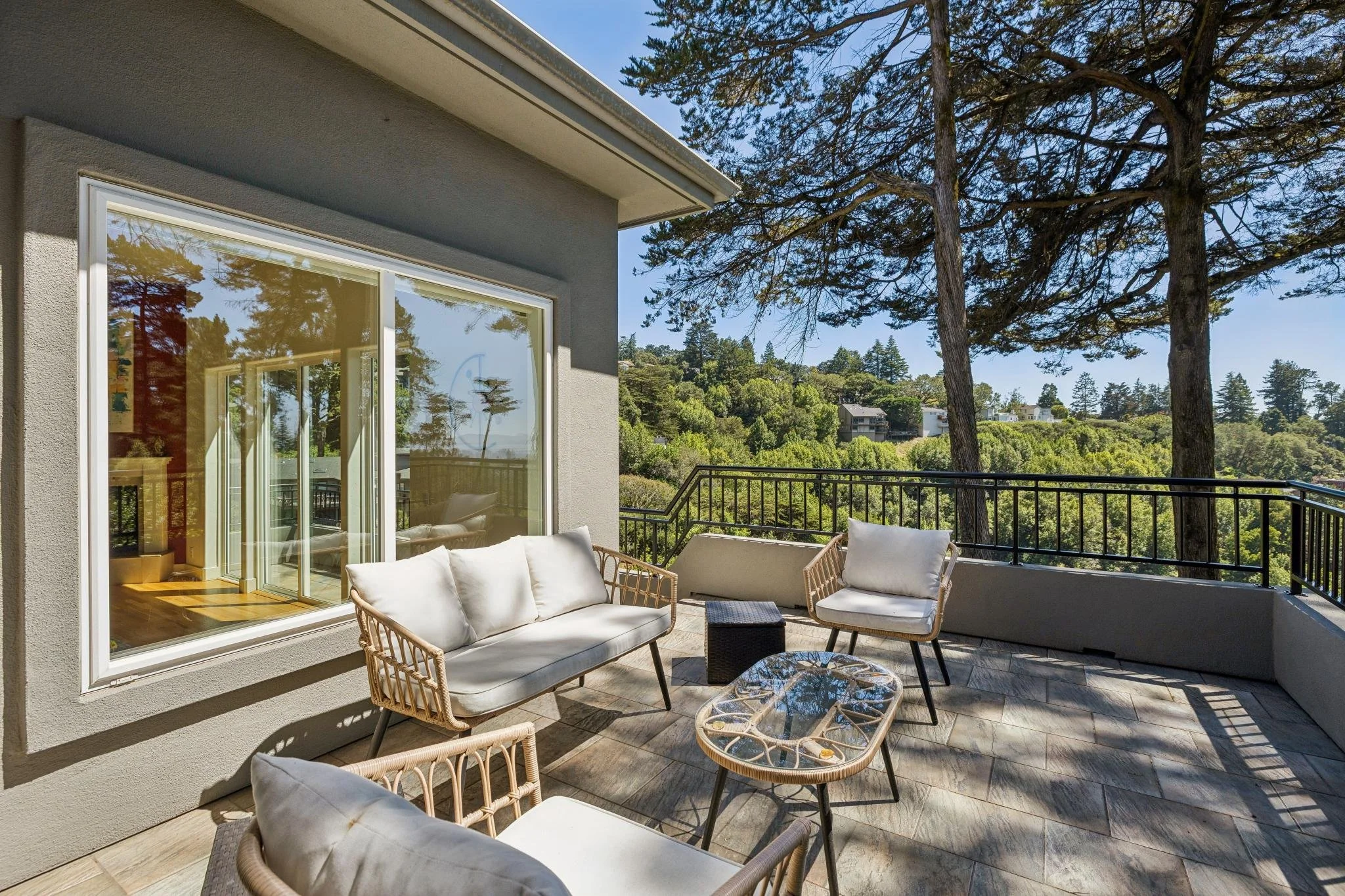 Balcony patio with outdoor seating, including a wicker sofa with white cushions, a wicker chair with white cushions, and glass-top tables, overlooking a lush green hillside with trees and houses in the distance.