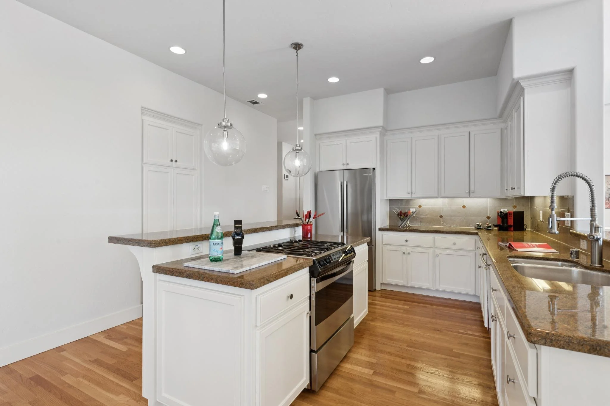 A modern kitchen with white cabinets, a granite countertop, stainless steel appliances, and hardwood floors, illuminated by ceiling lights and pendant lights.