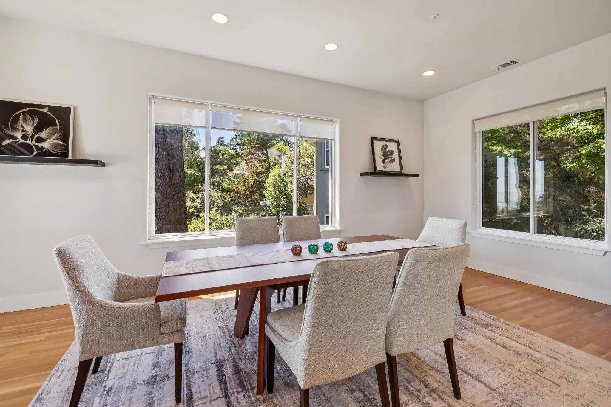 A bright dining room with a wooden table and six beige upholstered chairs, large windows with a view of greenery, minimalist black and white artwork on the walls, colorful candle holders on the table, and hardwood flooring.