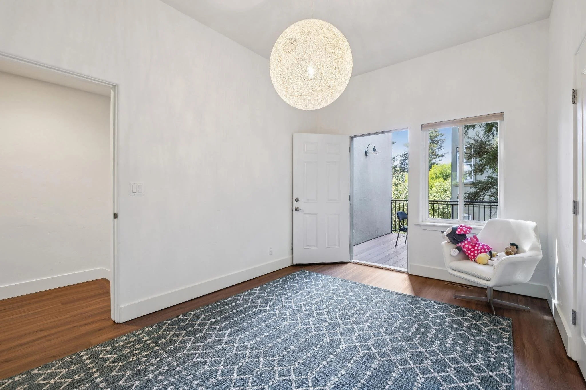 View of a bright, minimal living room with white walls, hardwood floor, large window, open door to balcony, and a white chair with stuffed toys on it.