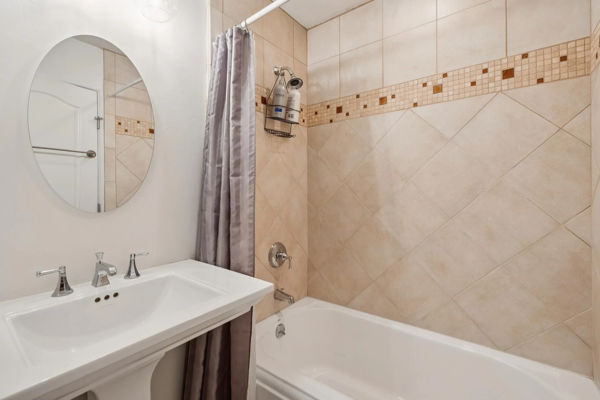 Bathroom with beige tiled walls, a white sink with silver fixtures, a large oval mirror, a shower area with a curtain, and a wire shelf holding shampoo and other bottles.