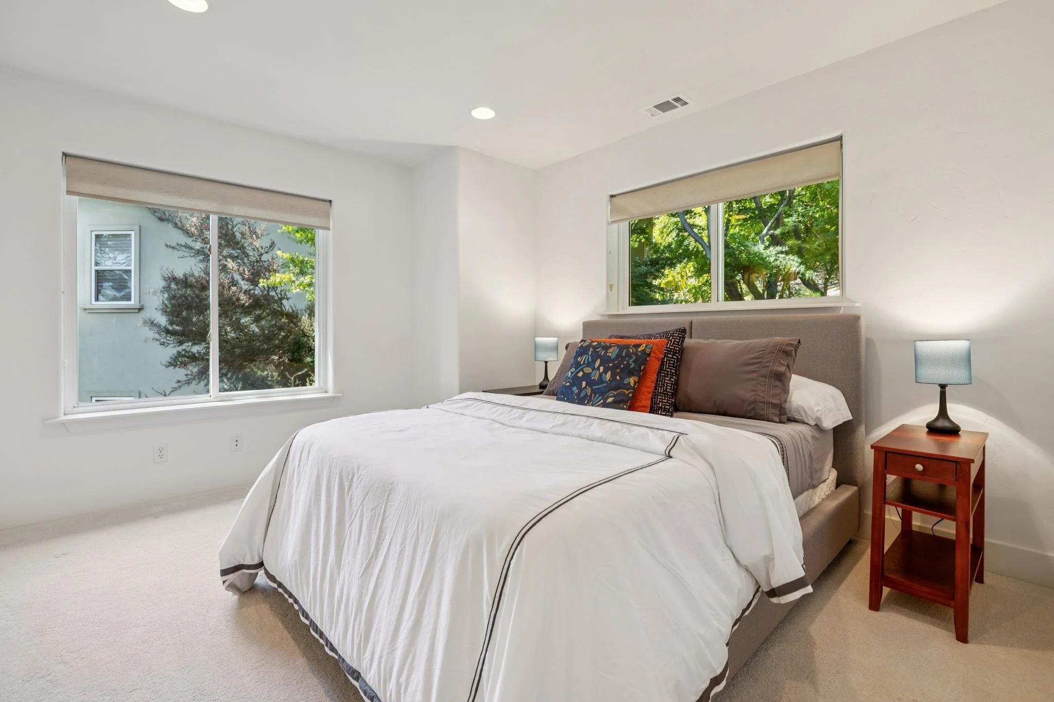 A bedroom with a large bed, beige bed frame, white bedding with dark trim, and multiple pillows including one with a colorful floral design. There are two bedside lamps on wooden nightstands, large windows showing green trees outside, white walls, an