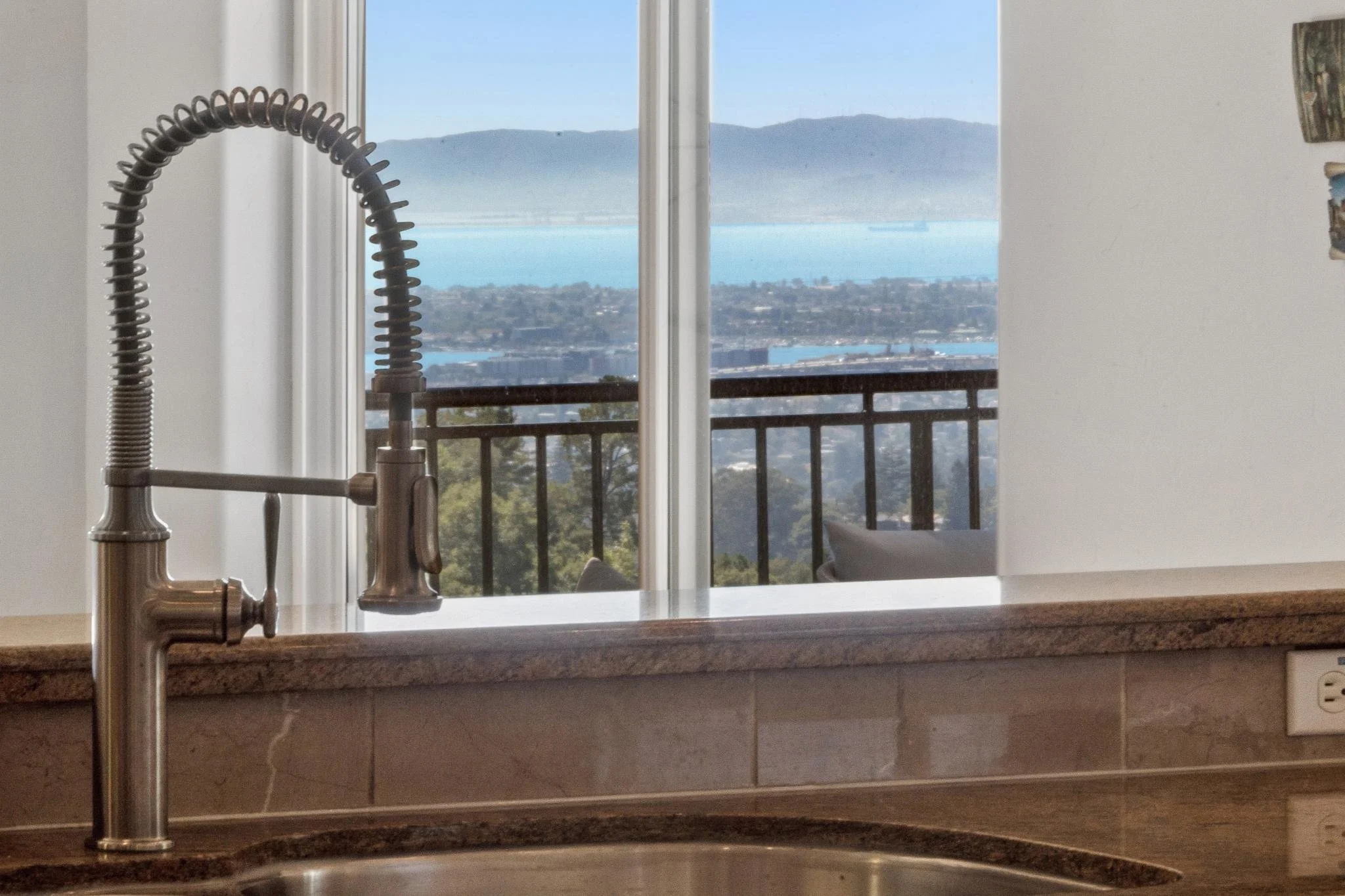 A kitchen sink with a modern gooseneck faucet, looking out a window onto a scenic view of a body of water, mountains, and a cityscape in the distance.