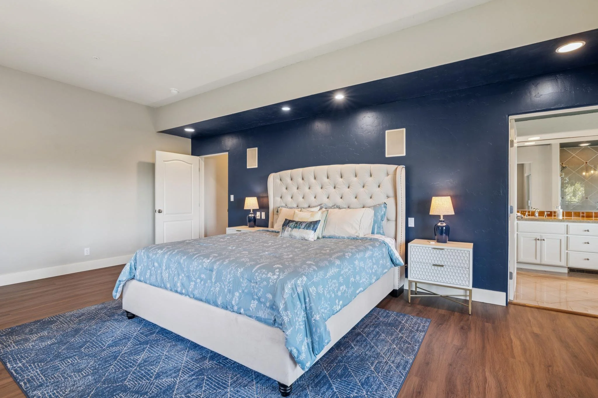 A bedroom with a white upholstered bed, blue and white bedding, two bedside tables with lamps, hardwood floors, blue area rug, and a blue accent wall with small wall panels. An open door leads to another room.