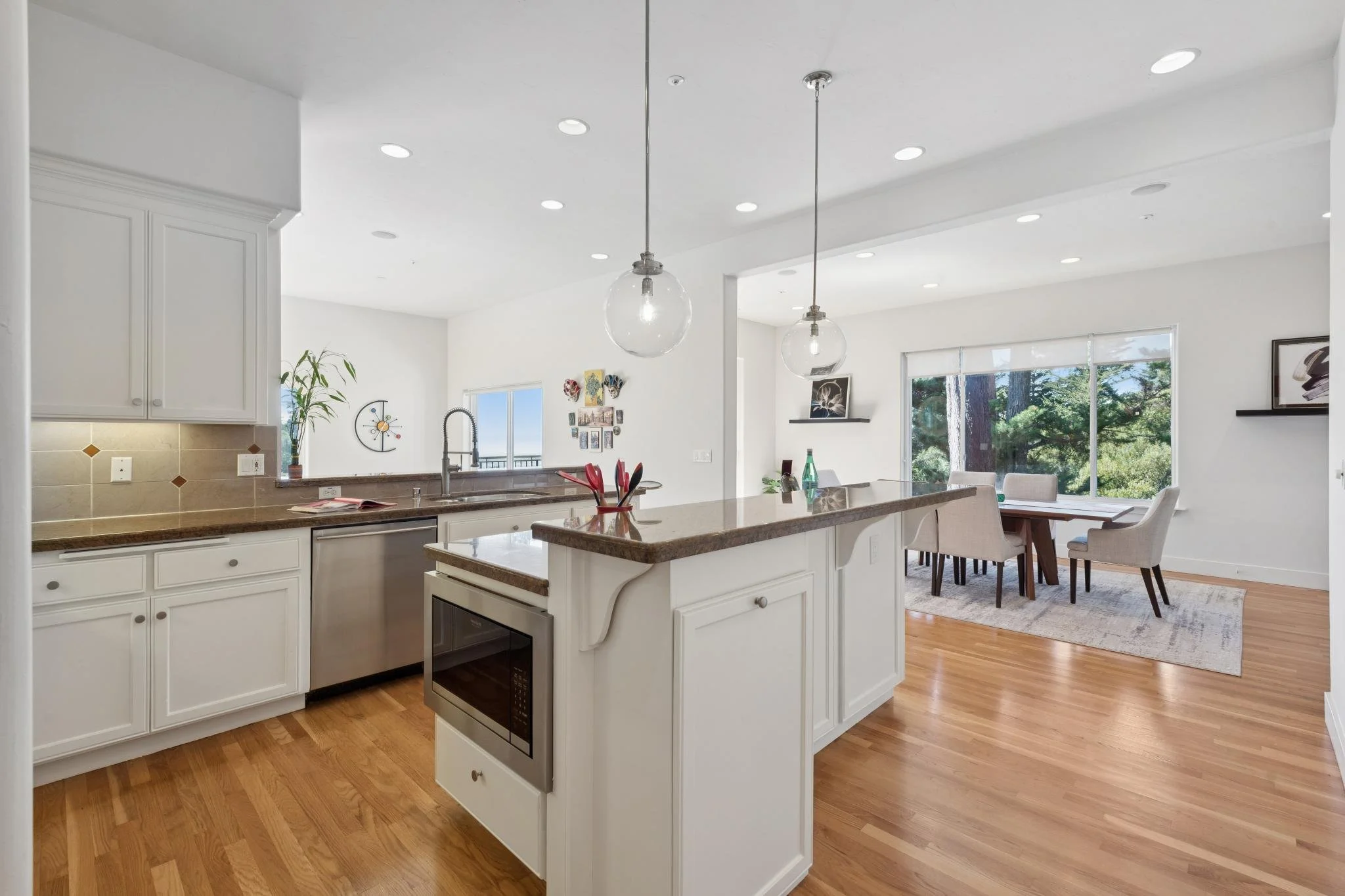Bright open-concept kitchen and dining area with white cabinetry, granite countertops, hardwood floors, large windows, and modern hanging pendant lights.