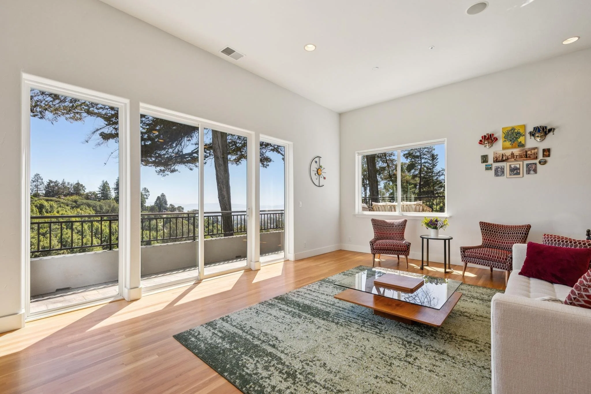 Living room with large windows showing trees and sky outside, beige furniture with patterned red and white cushions, clock on white wall, decorative wall art, wooden coffee table, and green patterned area rug.