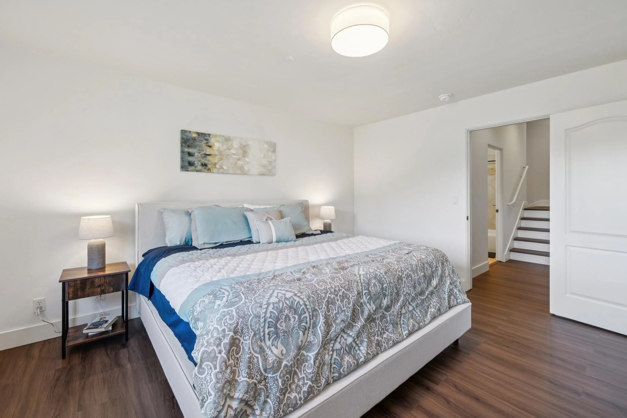 A bedroom with a large bed covered with a patterned quilt and multiple pillows, two bedside tables with lamps, a painting on the wall, and a doorway leading to stairs.