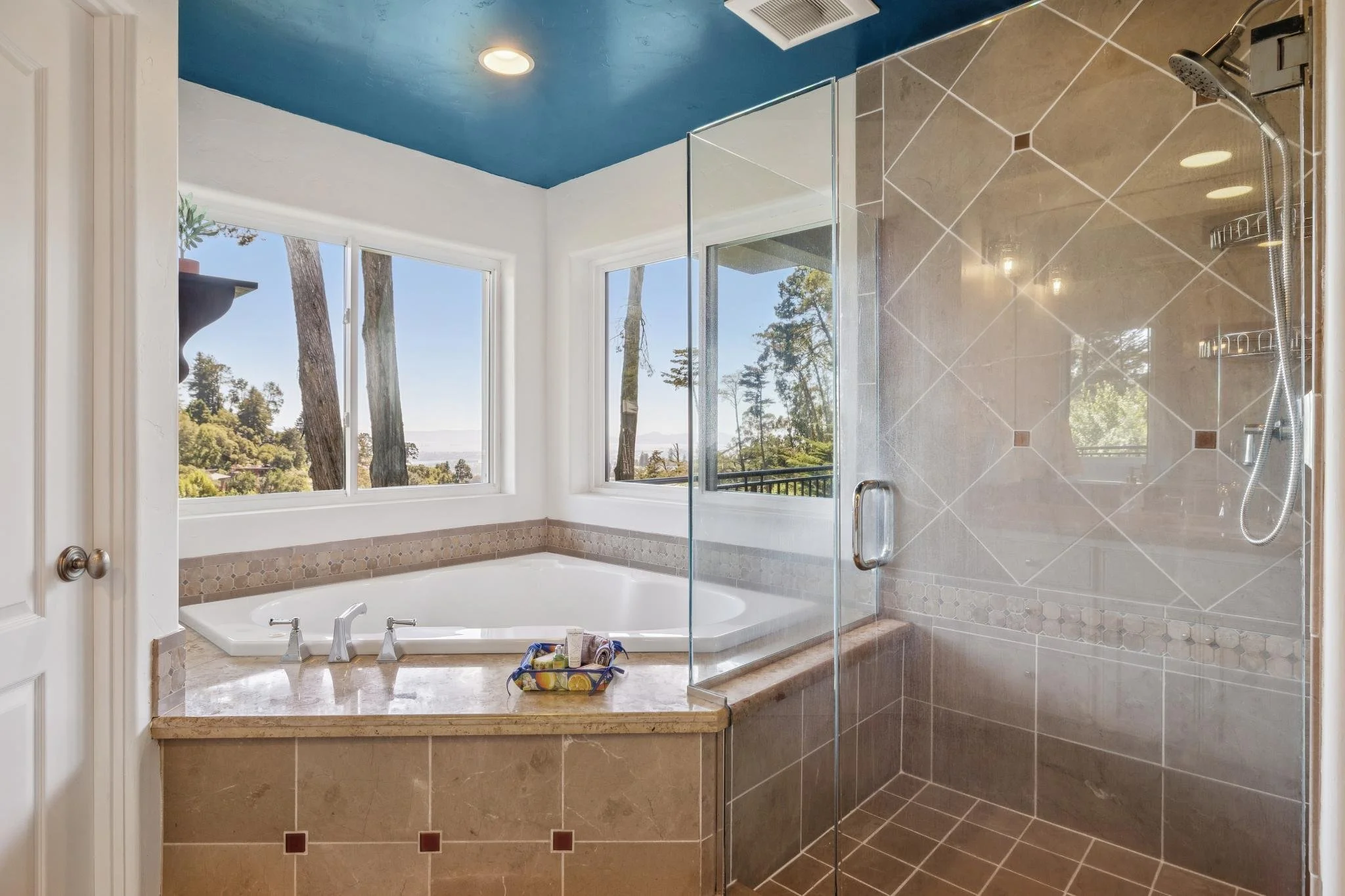 Bathroom with a large window showing trees outside, a bathtub with a bucket of toiletries, and a walk-in shower with glass enclosure, beige tiles, and a showerhead.