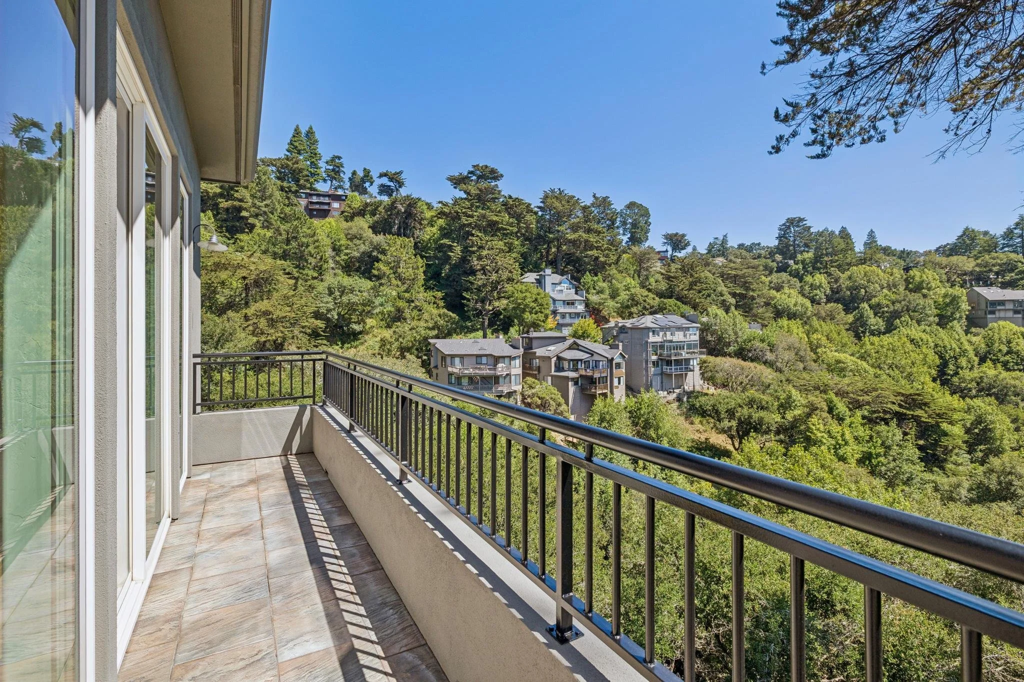 Balcony with tiled floor, white walls, black metal railing, overlooking a hillside neighborhood with trees and houses under a clear blue sky.