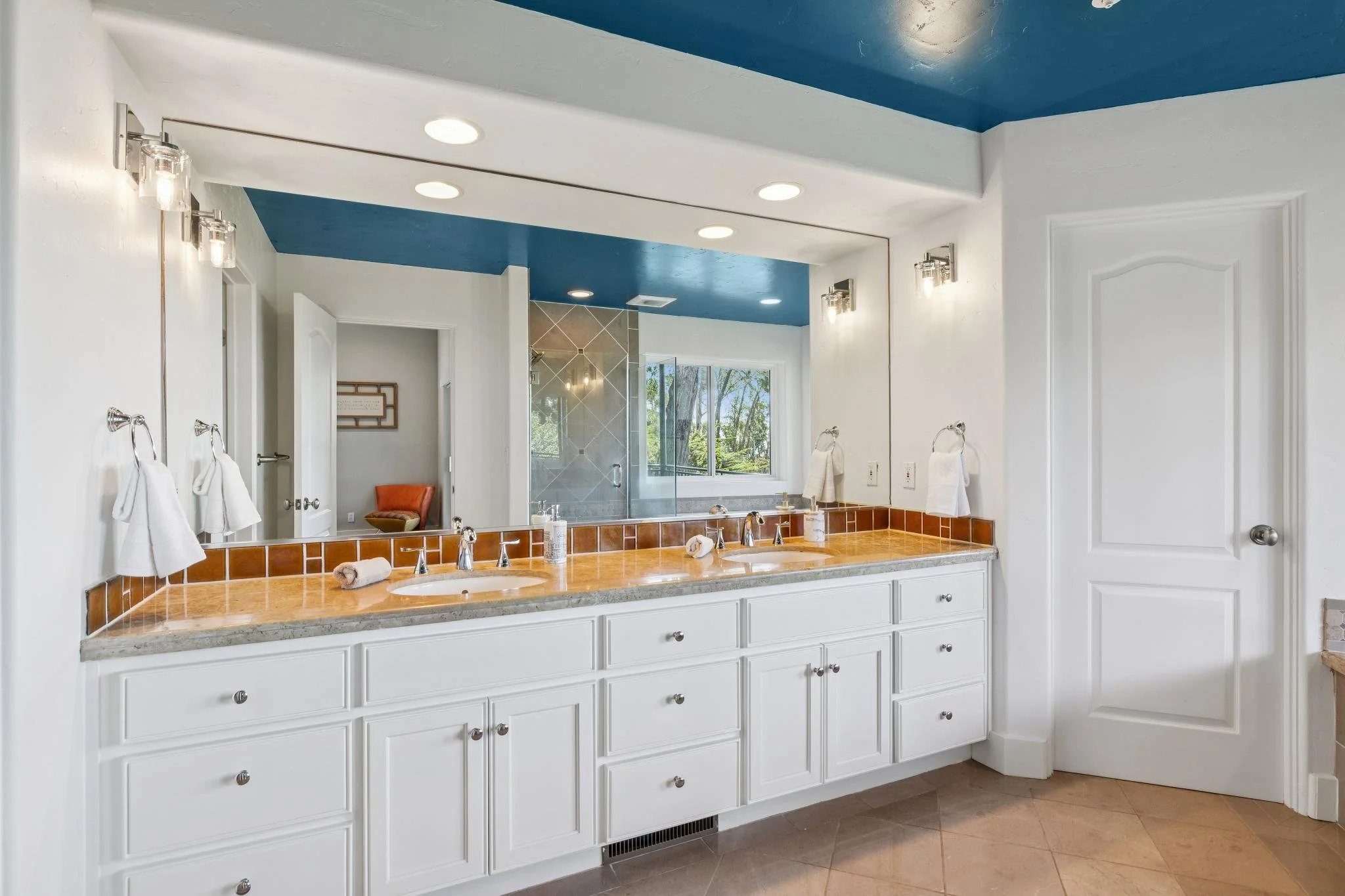 Bathroom with white cabinets, double sinks with tan tile countertops, large mirror, three wall-mounted light fixtures, towel rings with towels, and a window showing trees outside.