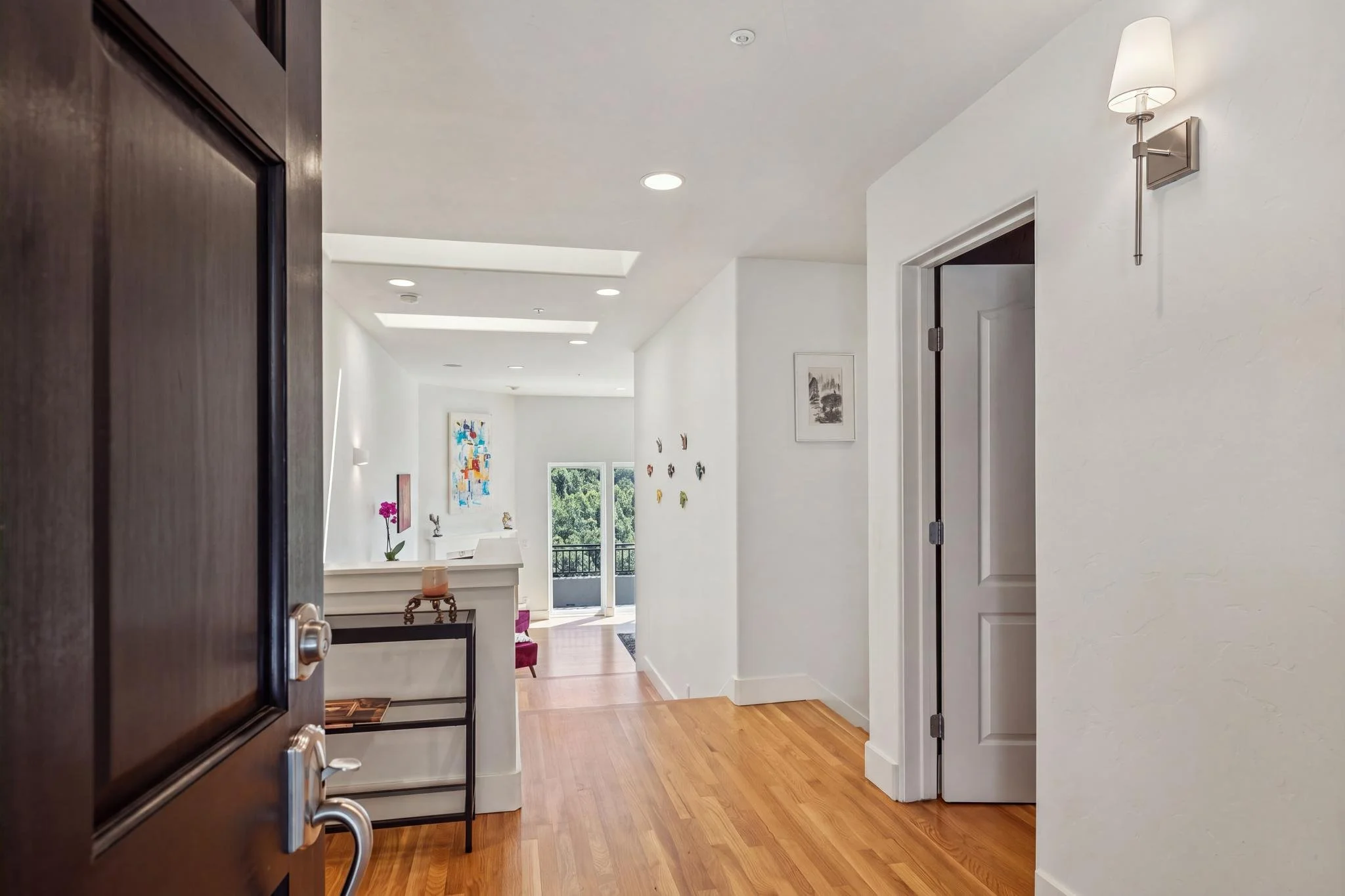 Interior view of a modern home living space seen through an open door, featuring white walls, hardwood floors, artwork, a table with a pink orchid, and a balcony with greenery outside.