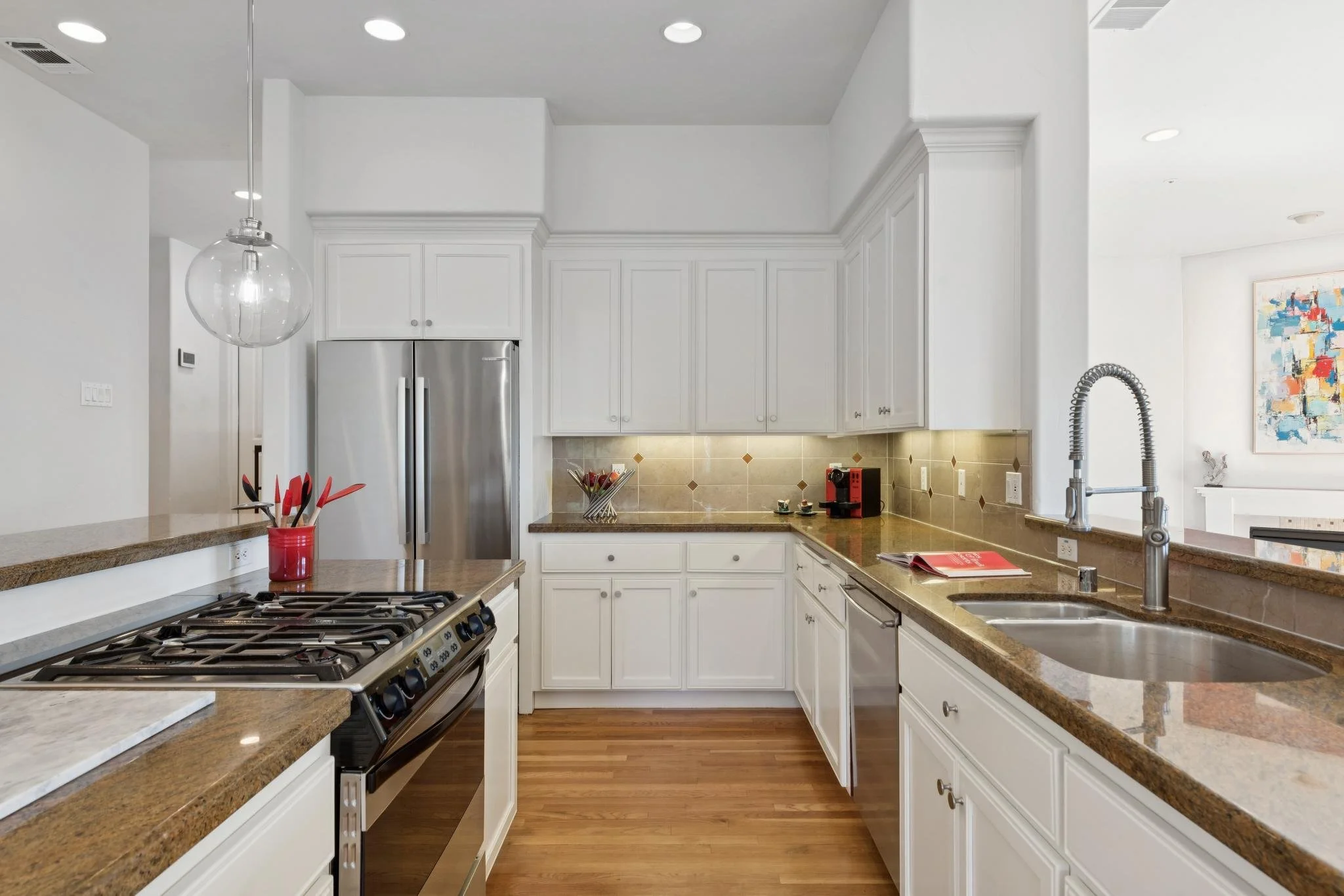A modern kitchen with white cabinets, brown granite countertops, a stainless steel refrigerator, a gas stove, a sink with a tall faucet, and colorful artwork on the wall.