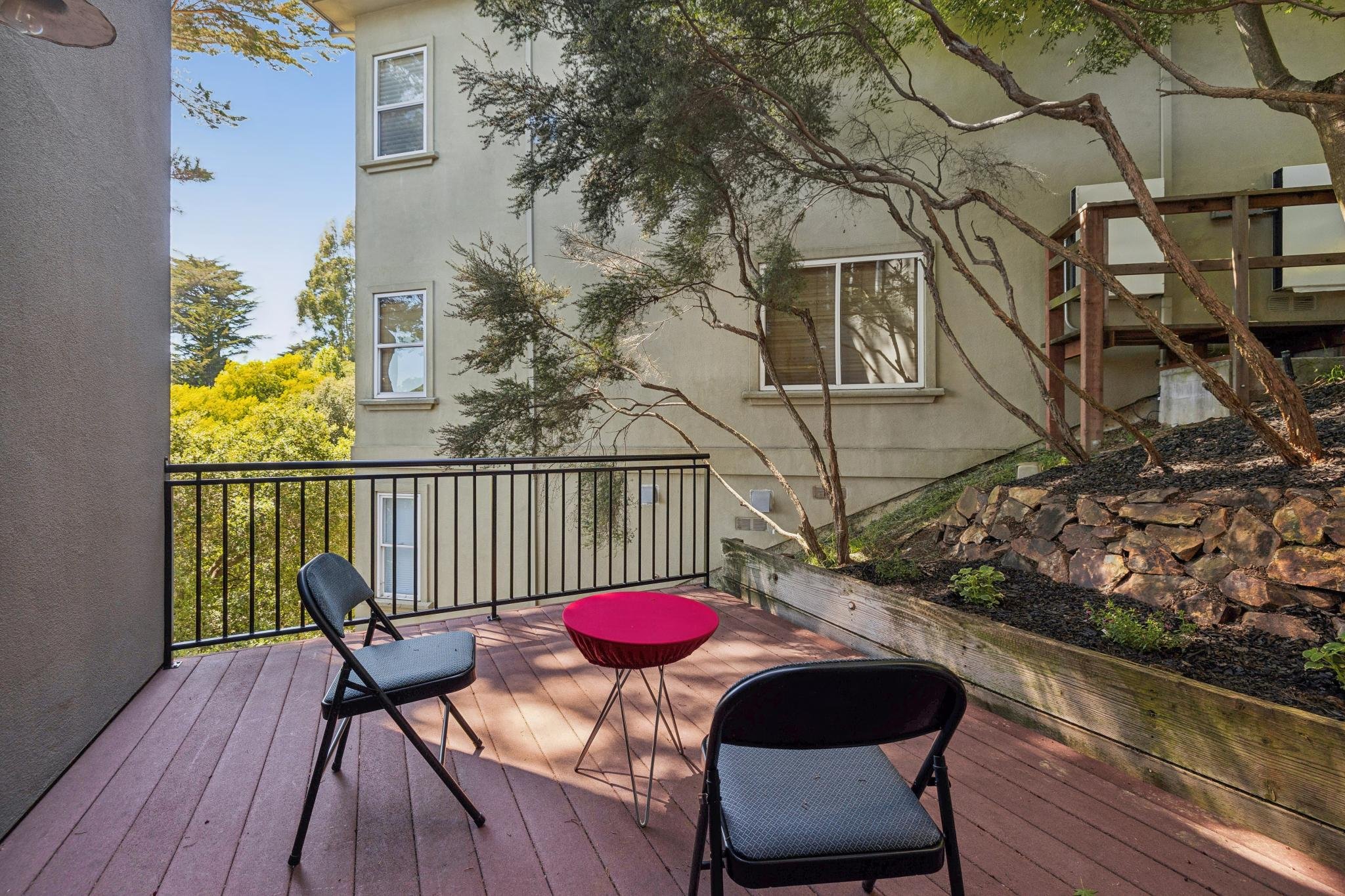 A cozy backyard patio with patio furniture, including a round red table and three black chairs on wooden decking, enclosed by a black metal railing, with a lush landscape, a small tree, and a building with multiple windows in the background.