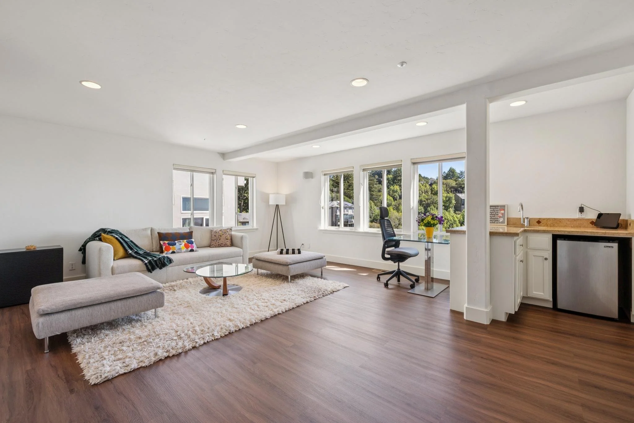 A spacious living room with white walls, several large windows, a beige sofa with colorful pillows, a white shaggy rug, a small ottoman, a round coffee table, a standing lamp, a black office chair, and a kitchen area with a granite countertop and cab