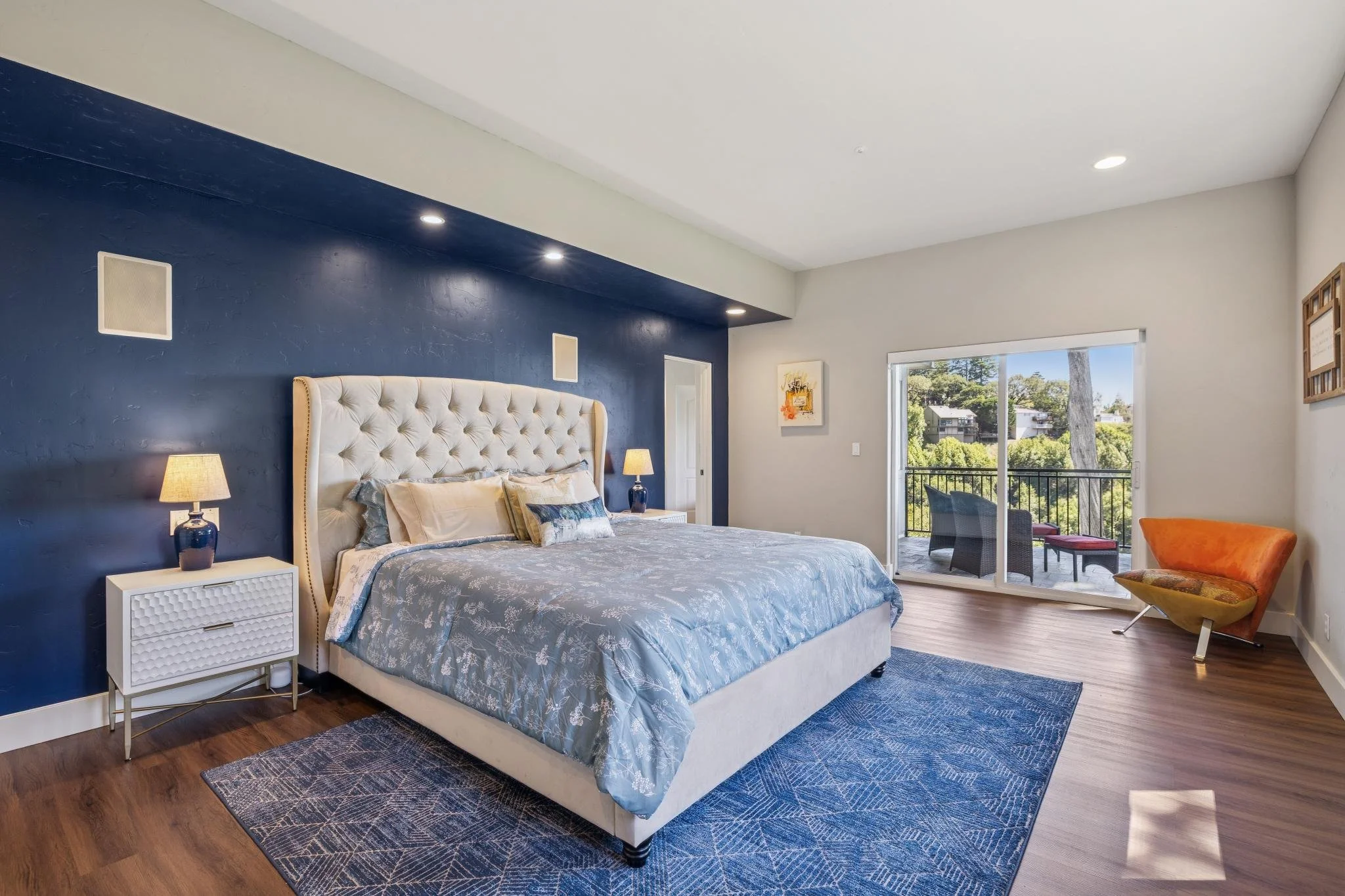 A bedroom with blue and neutral-colored walls, a large upholstered headboard, side tables with lamps, a sliding glass door leading to a balcony, and various chairs and artwork.