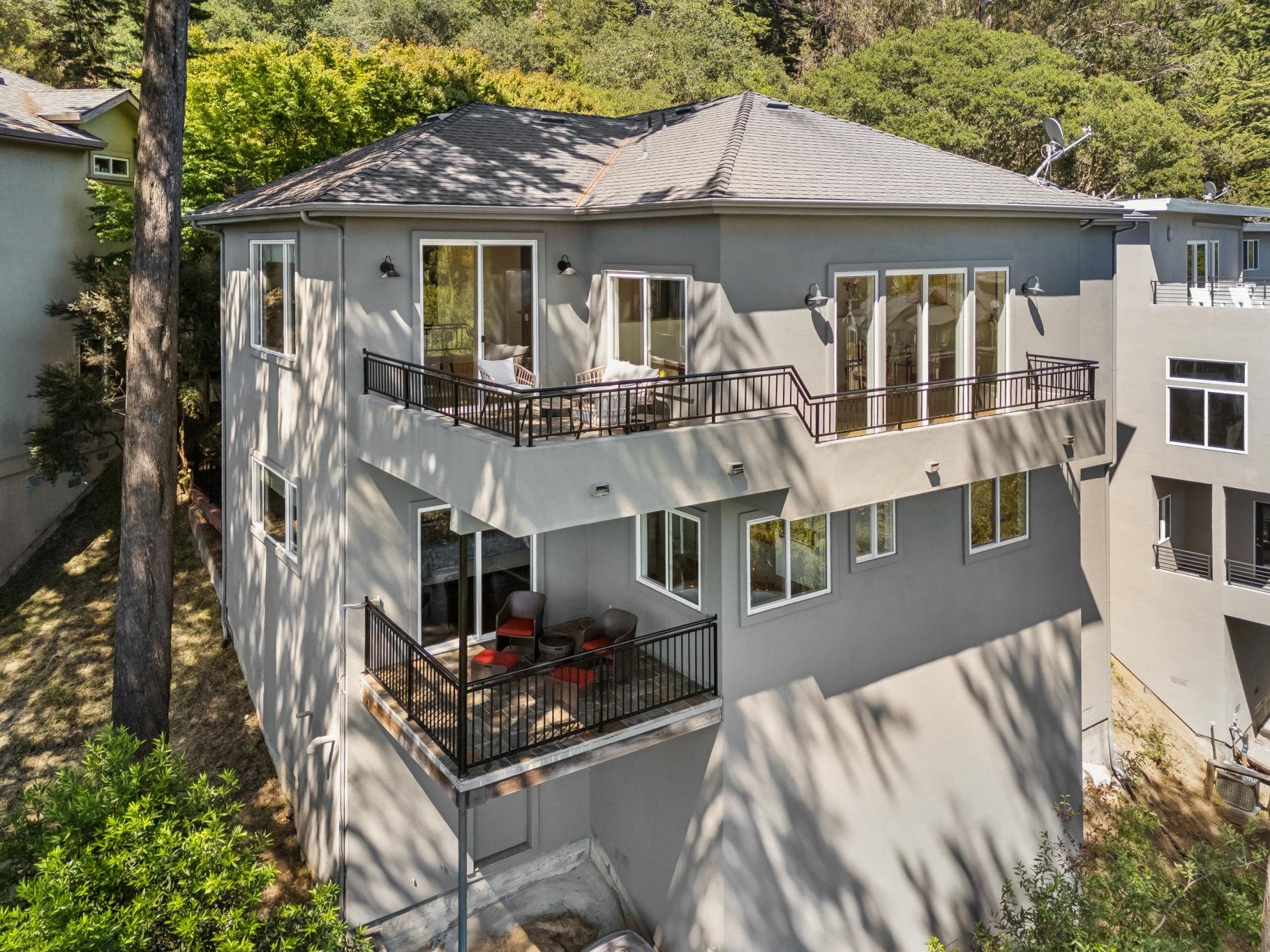 A multi-story residential building with multiple balconies overlooking a green, wooded area. The balconies are furnished with outdoor seating and are accessed by glass sliding doors. The building has a modern style with light gray exterior walls and black railings. Satellite dishes are visible on the roof.