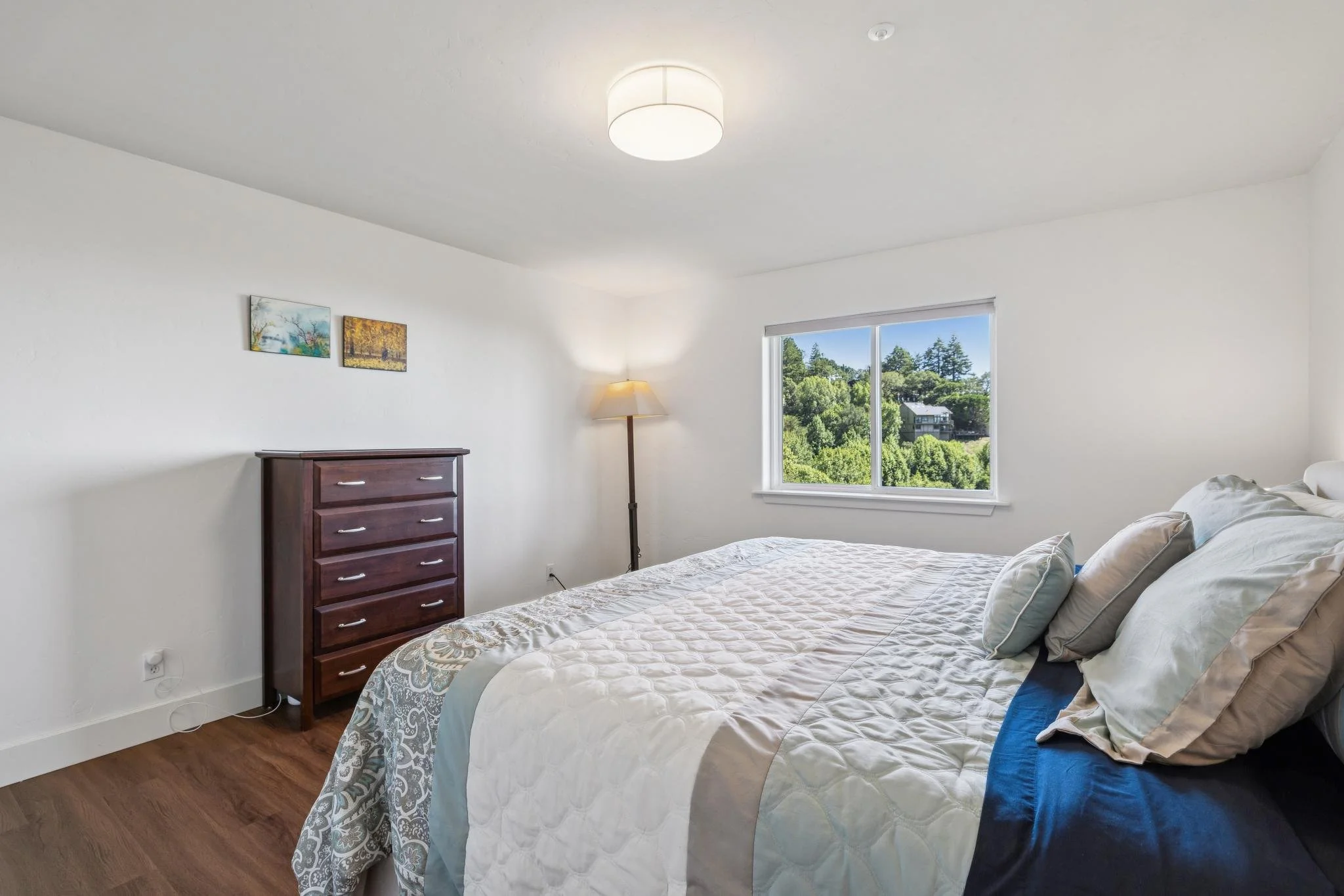 Bright bedroom with white walls, large window showing trees outside, wooden floor, bed with white and beige bedding, pillows, furnished with a dresser and standing lamp, ceiling light.