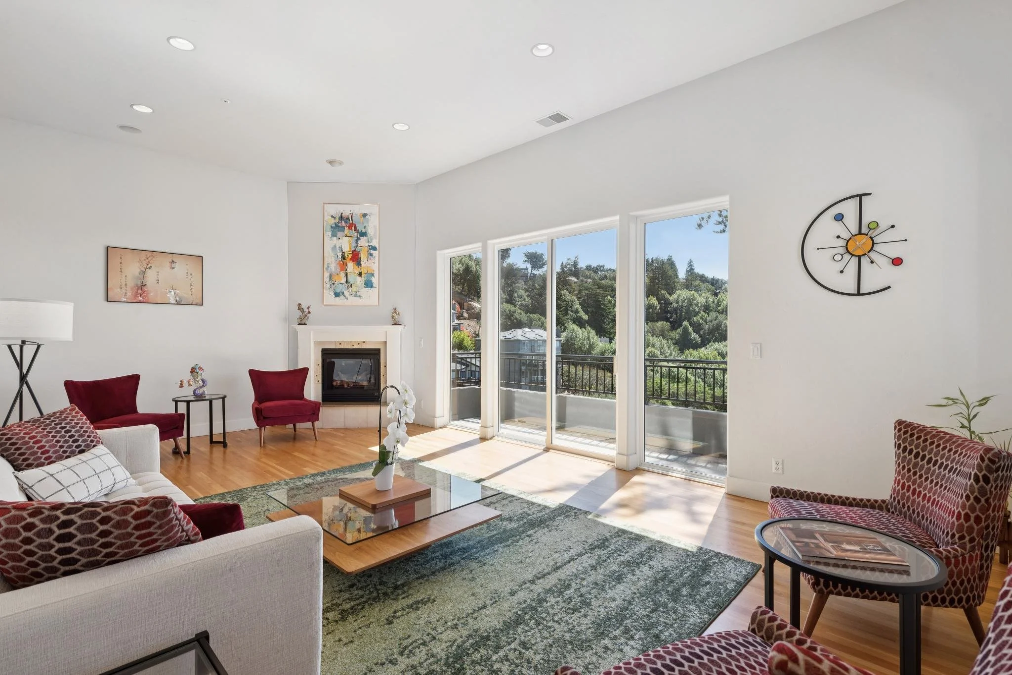 Bright living room with white walls, large windows showing green trees and blue sky, various red and patterned armchairs, a white sofa with patterned pillows, a glass coffee table with a flower vase, wall art, a clock, and a fireplace.