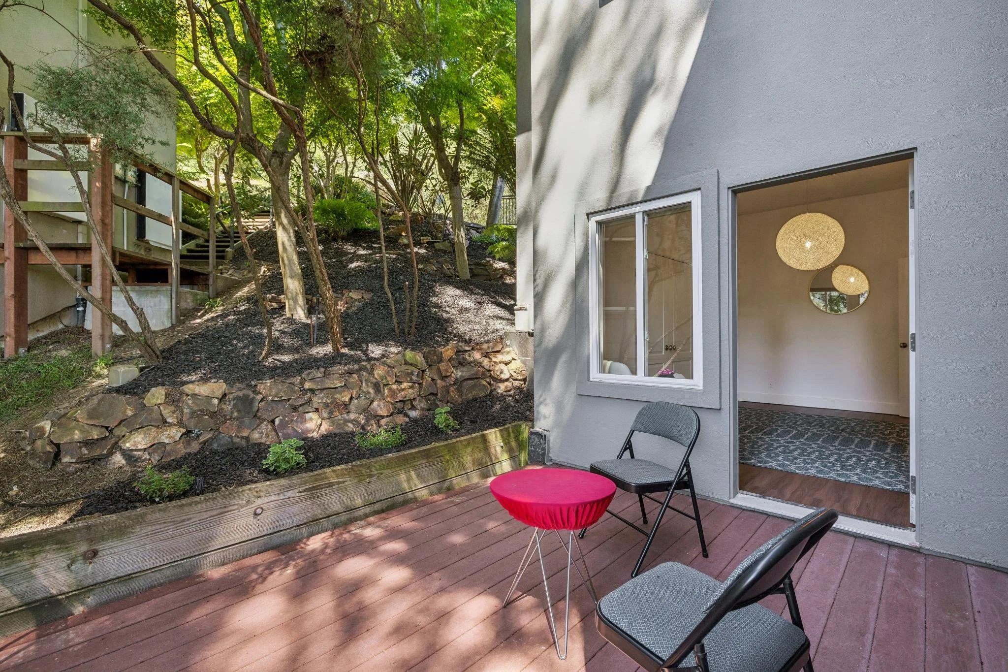 A backyard patio area with wooden decking, black chairs, and a small round red table. The patio is adjacent to a house with white walls, open sliding glass door, and interior lighting fixtures. Behind the patio is a garden with greenery, trees, and r