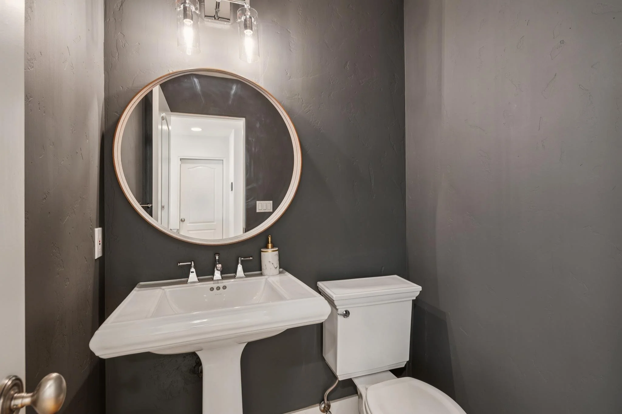 A modern bathroom with a dark gray textured wall, a white vanity with a sink, a round mirror, a small cabinet, and a toilet.