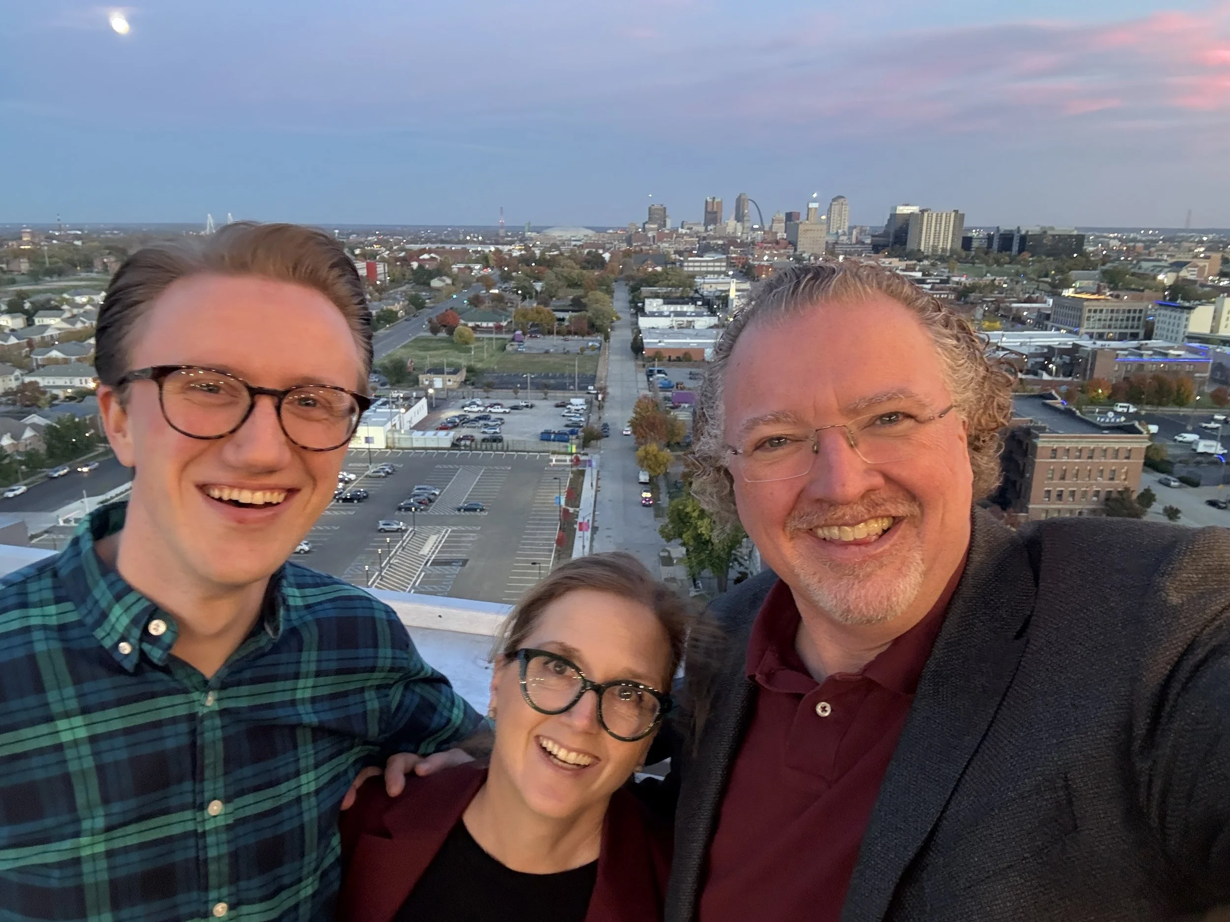 Four smiling people taking a selfie on a rooftop with a city skyline and a partly cloudy sky in the background.