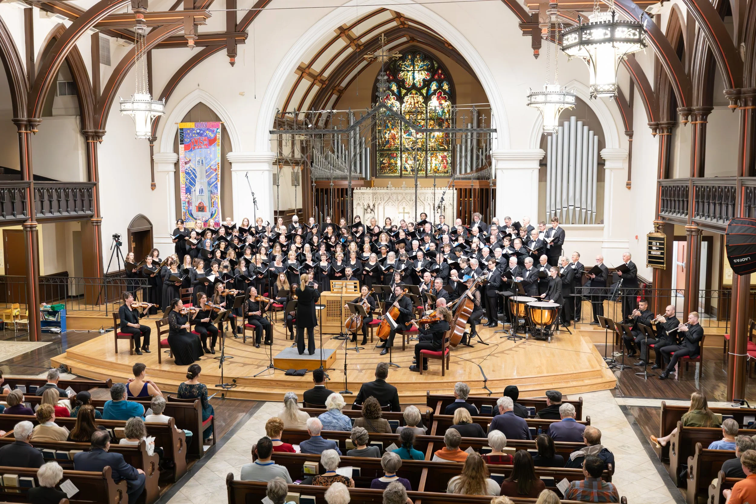 Erin Freeman conducts a chorus and orchestra inside a church with stained glass windows and wooden architectural details, as an audience watches.