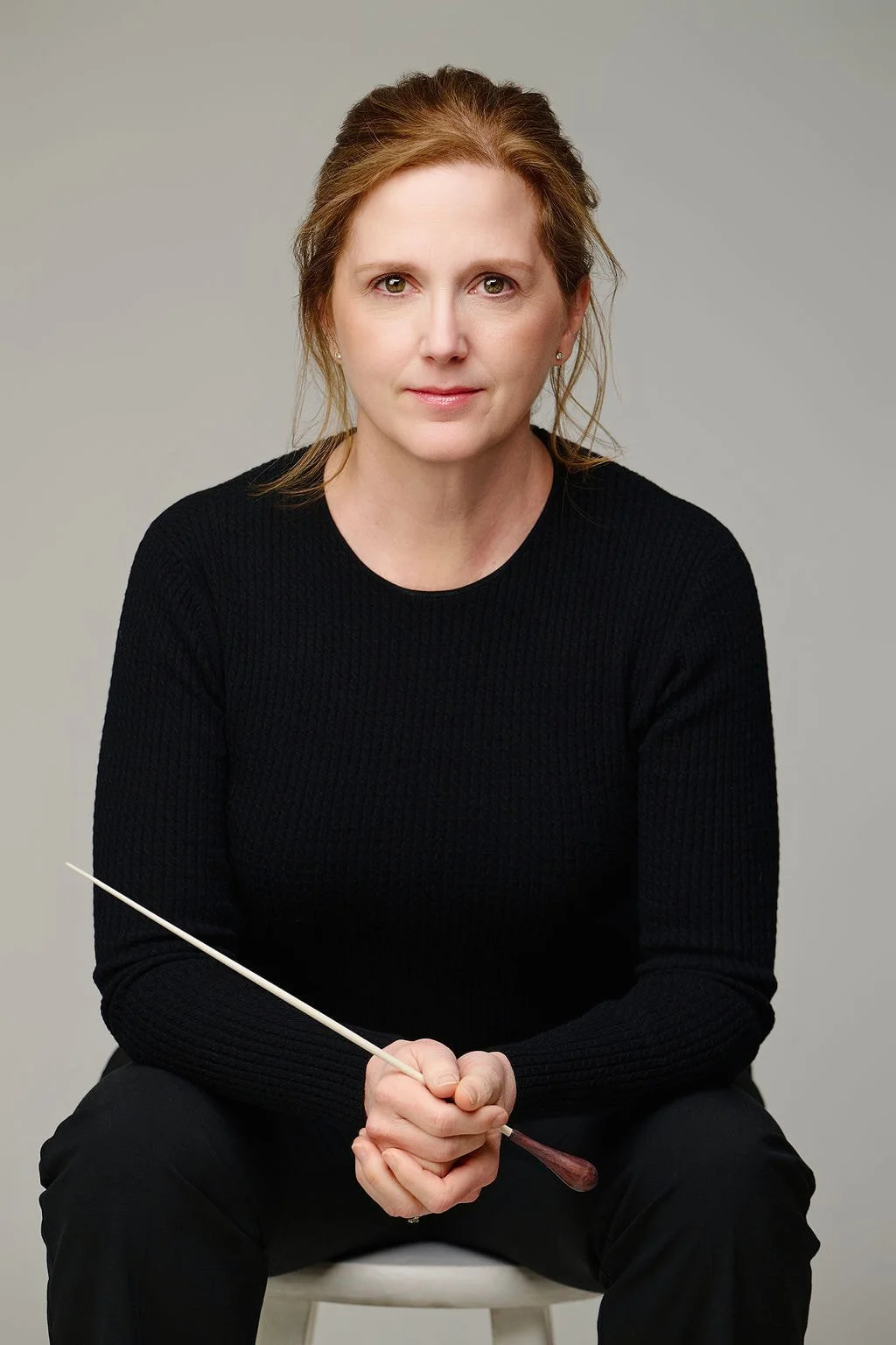 A woman with light skin and brown hair, wearing a black sweater, sitting on a white stool, holding a conductor's baton.