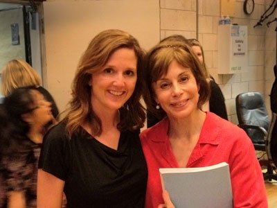 Two women smiling indoors, one holding a folded paper or document.