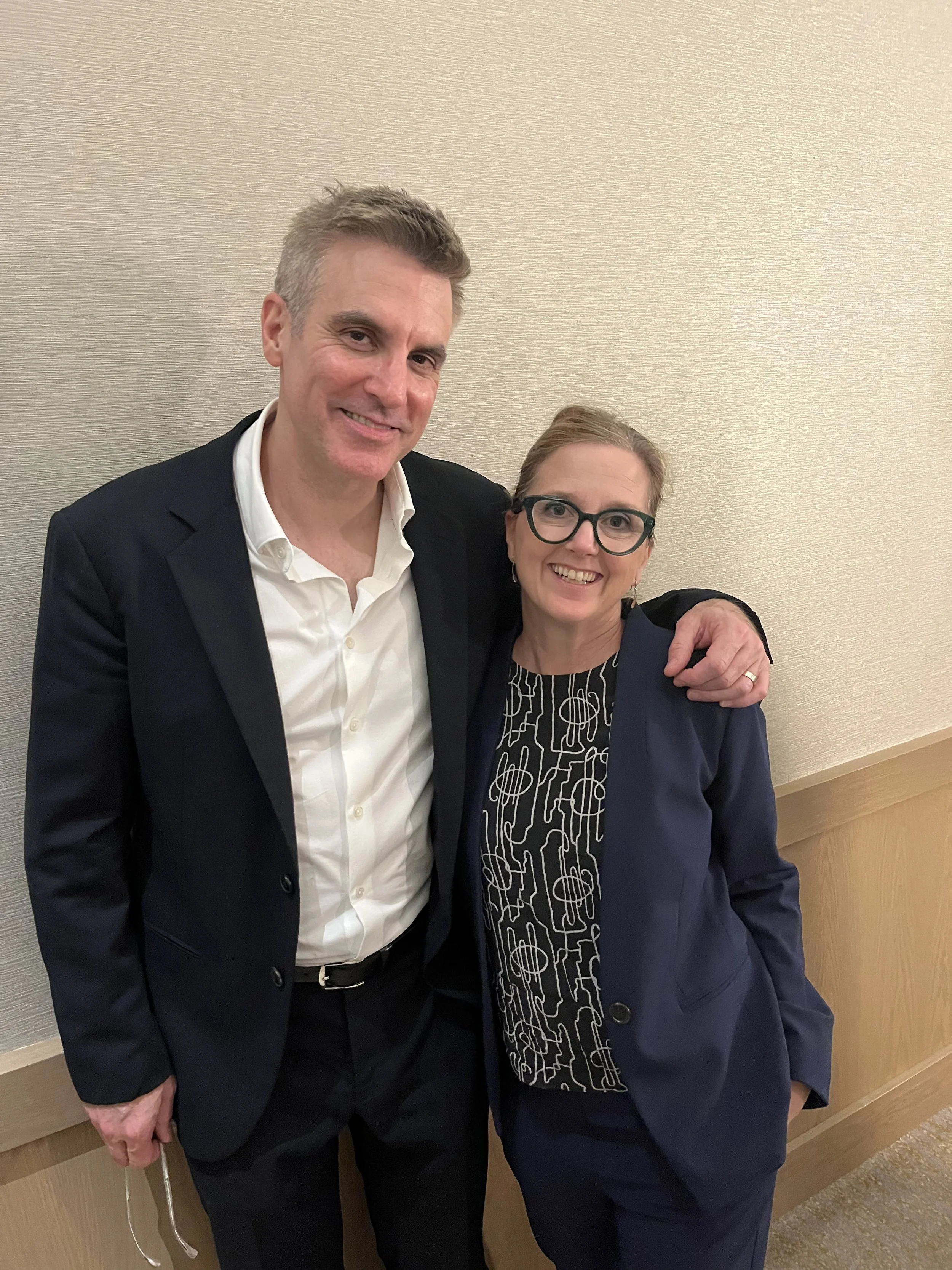 A man and a woman are standing together indoors, smiling. The man is wearing a white dress shirt and a black blazer, and the woman is wearing glasses and a dark blazer over a patterned top. They are positioned close to each other with the woman's arm
