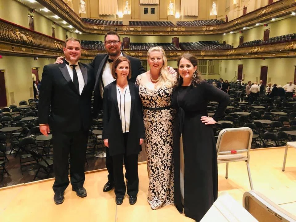 Five people dressed in formal attire standing on a stage inside a theater with empty seats in the background.