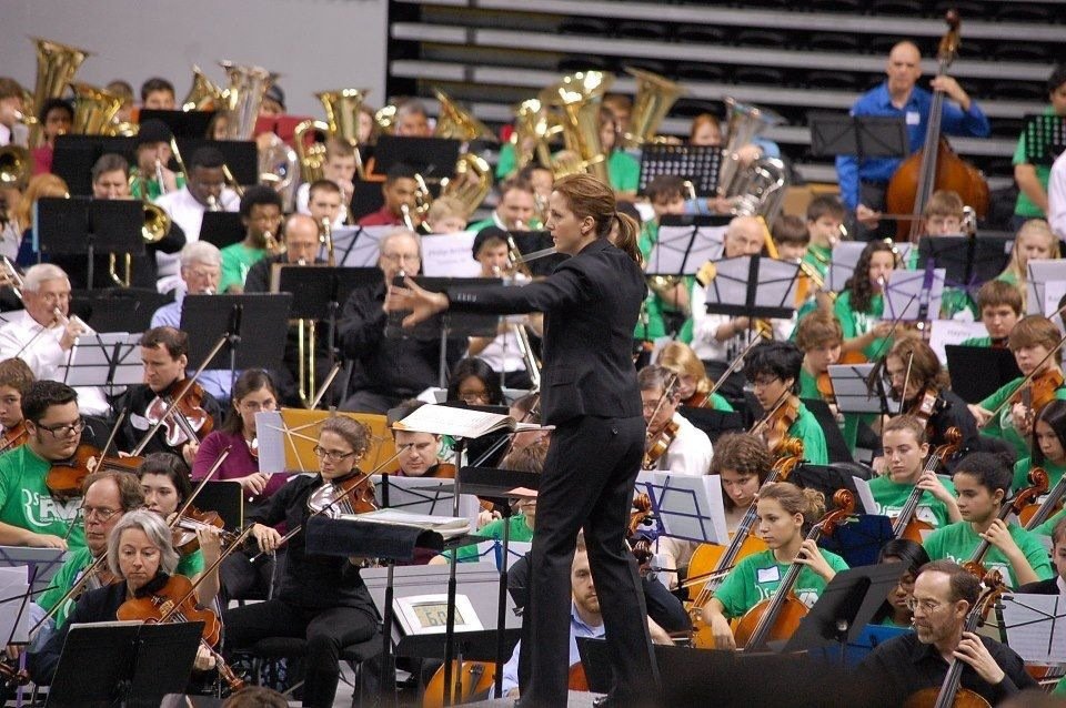A symphony orchestra performing on stage, led by a conductor. The orchestra consists of musicians playing violins, cellos, and brass instruments. The musicians are dressed in green, purple, and black, and the conductor is in a black suit, facing the 